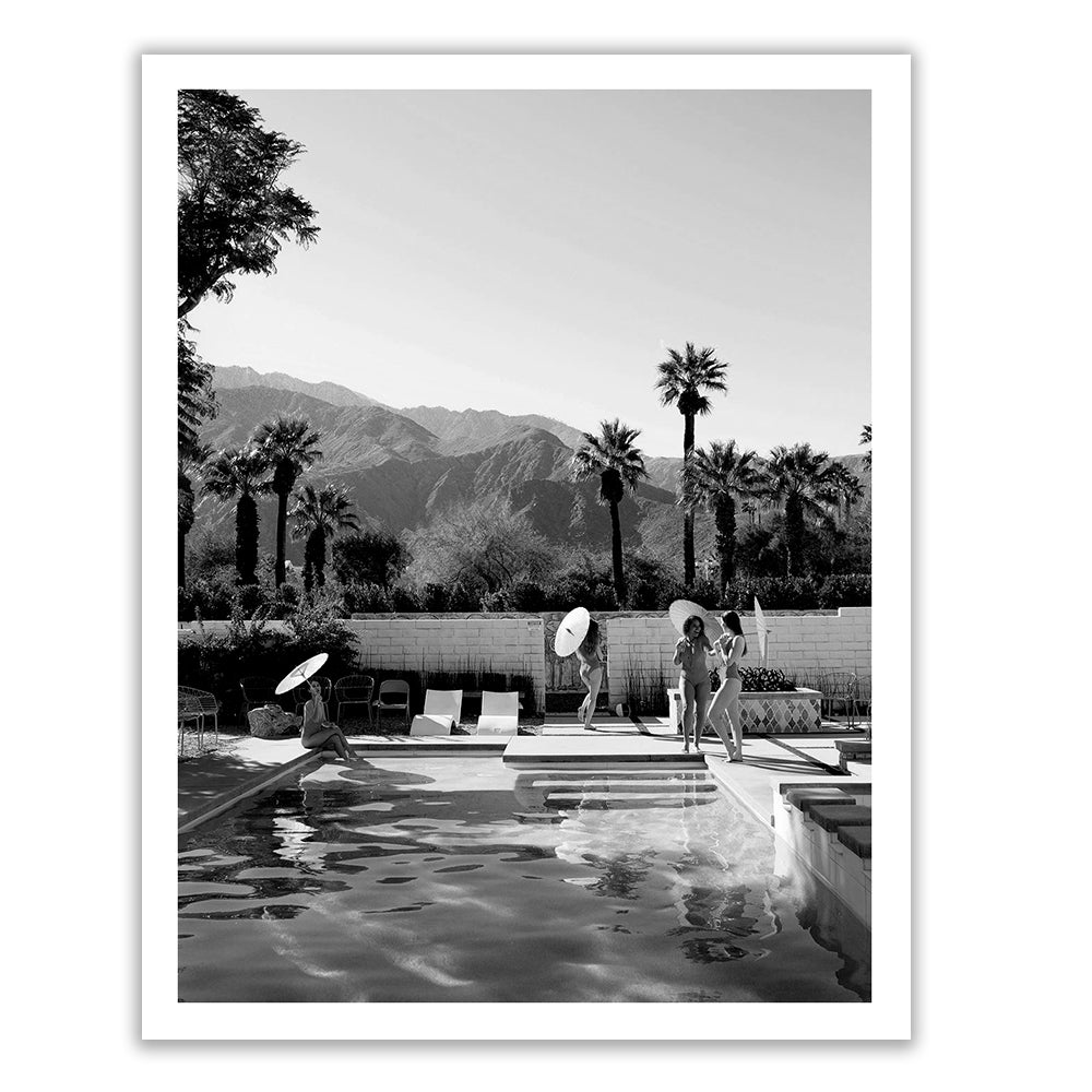 The black-and-white "Poolside Mermaids B&W" photo by Francesca Owen captures a serene swimming pool scene with people around it. Two individuals are holding parasols near the pool, with mountains and palm trees in the background. This limited edition print is created using archival pigments, ensuring lasting quality and detail.