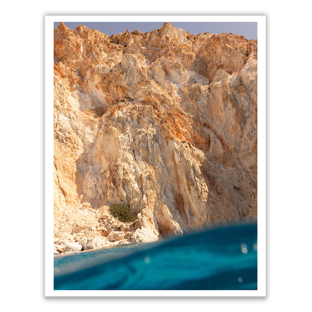 In "Magic in Milos #7" by Francesca Owen, a towering cliff of rugged, colorful rock formations rises above a bright blue water surface. Captured with archival pigments on fine art paper, the scene offers a dramatic contrast between the earthy tones of the cliff and the vivid ocean in the foreground, and it comes with a certificate of authenticity.