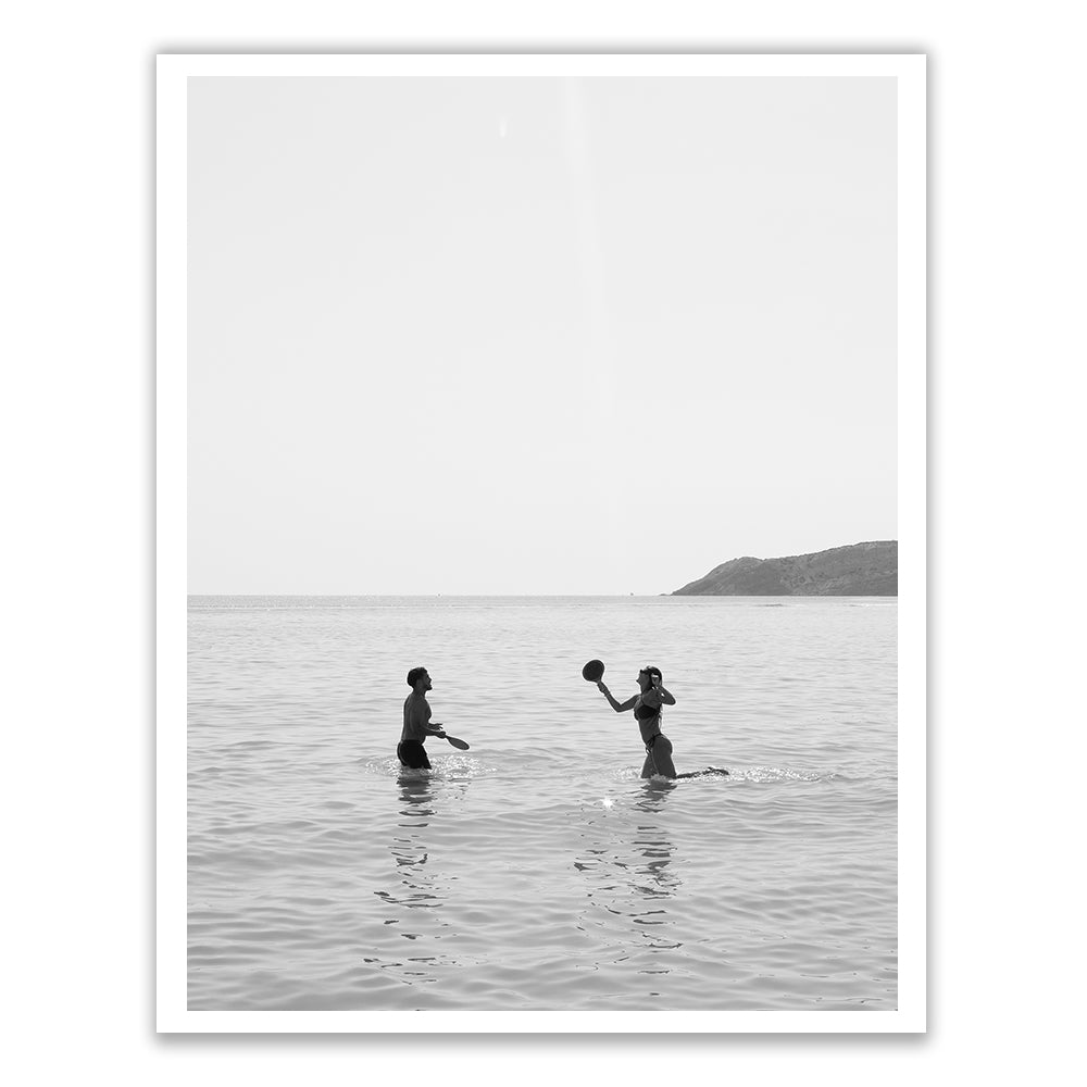 In the black and white photograph titled "Magic in Milos #8" by Francesca Owen, two people are seen playing with a ball while standing waist-deep in the ocean. The stunning backdrop of hills and clear skies adds to the scene’s timeless quality, evocatively captured using archival pigments on fine art paper.