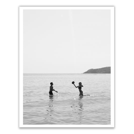 In the black and white photograph titled "Magic in Milos #8" by Francesca Owen, two people are seen playing with a ball while standing waist-deep in the ocean. The stunning backdrop of hills and clear skies adds to the scene’s timeless quality, evocatively captured using archival pigments on fine art paper.