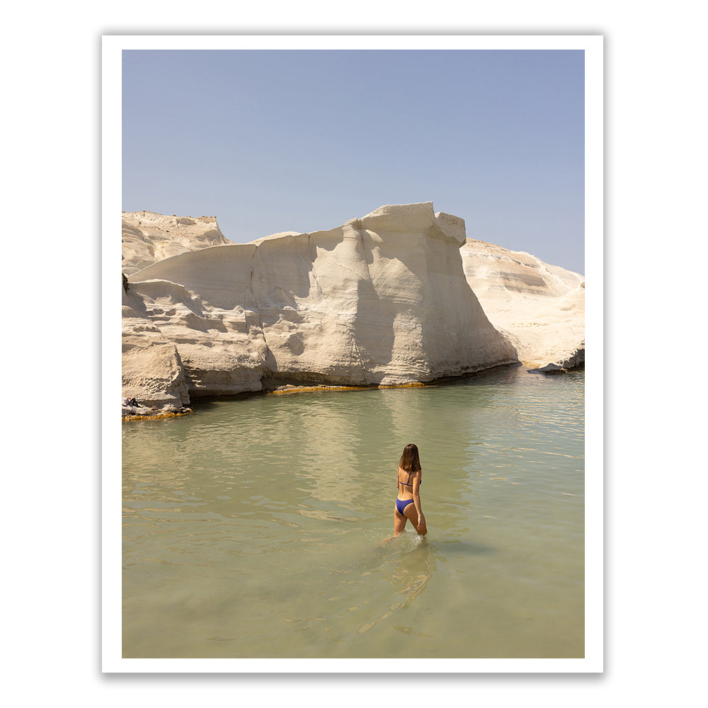 A person stands in shallow, clear water, facing large, light-colored rock formations under a sky as pristine as the archival pigments on Francesca Owen's "Magic in Milos #12.