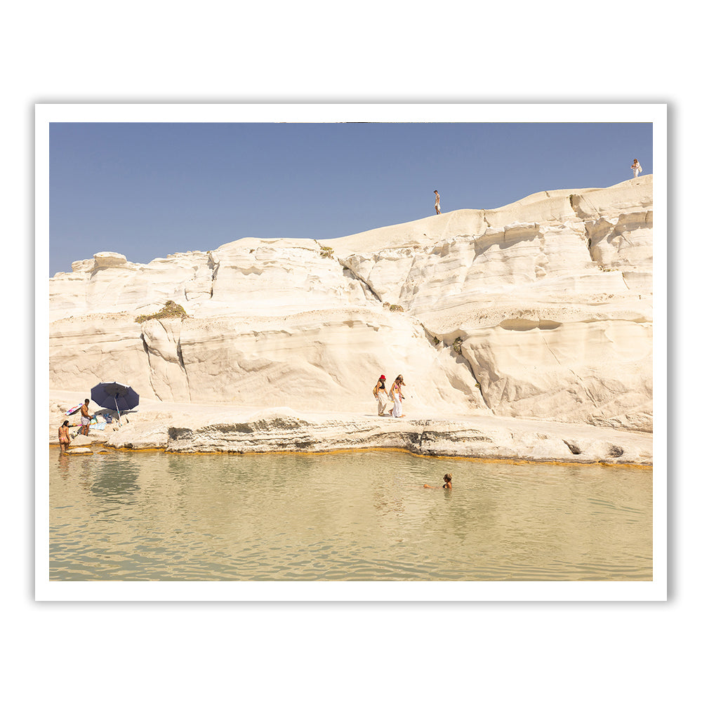 Visitors are captivated by Magic in Milos #13, a stunning scene by Francesca Owen that showcases people basking in the sun at a uniquely beautiful beach with eye-catching white rock formations. Some wander and lounge on the rocks while others swim in the crystal-clear water below. A blue umbrella offers shade near the water's edge, making this picturesque moment perfect for capturing with archival pigments.