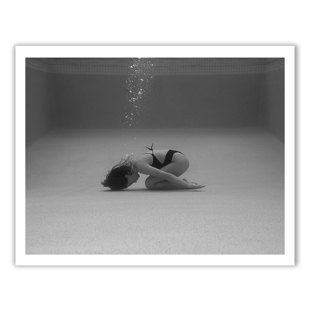 Self Reflection 15” by Francesca Owen is a black and white photograph depicting a person underwater, kneeling on the pool floor with their head bowed as bubbles ascend towards the surface. The pool walls form a visible backdrop. This image, printed on fine art paper, comes with a certificate of authenticity for collectors.