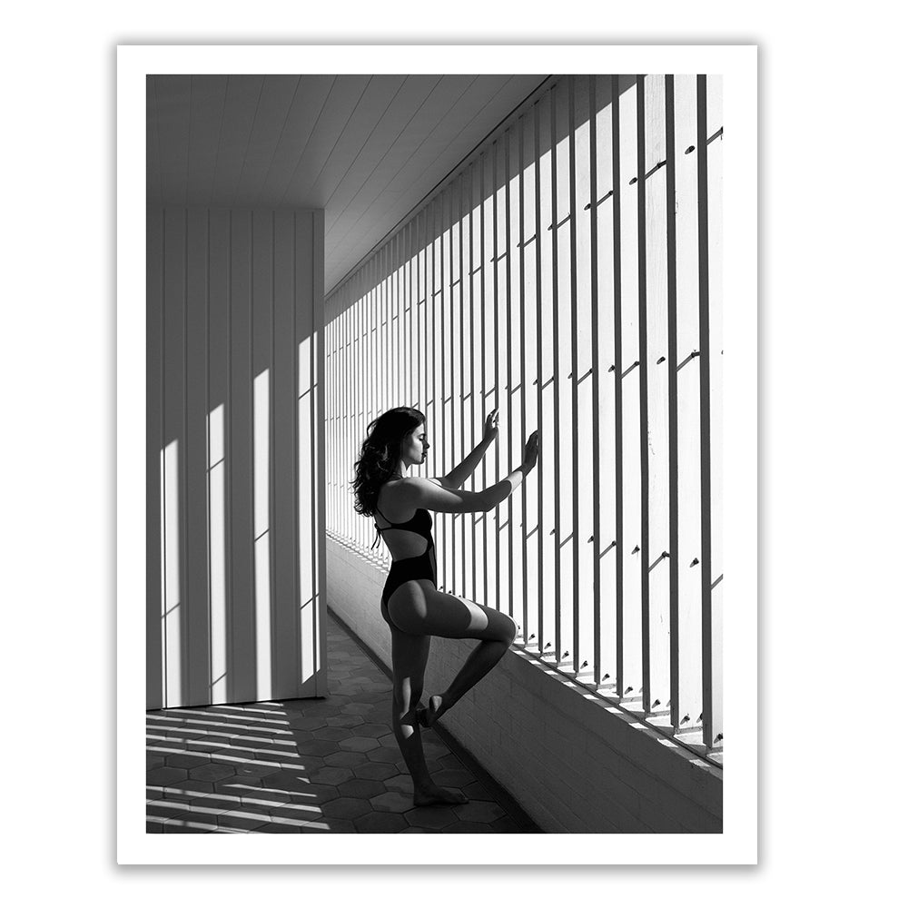 Self Reflection 09 by Francesca Owen features a black and white photo of a woman in a one-piece swimsuit standing by vertical bars casting shadows. She is looking out into the distance with one leg bent and an arm extended to touch the bars. This striking image, printed on fine art paper, is bathed in sunlight and deep shadows.