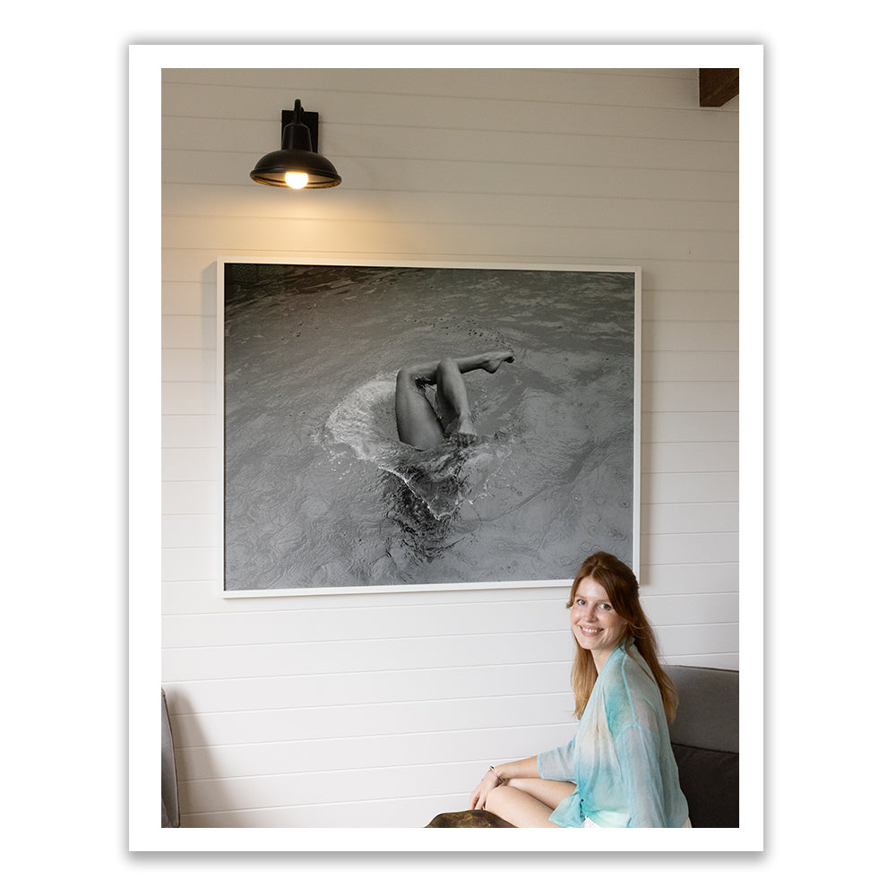 A woman sits on a couch in a modern room, smiling at the camera. Behind her, "Self Reflection," a large black and white photograph framed with archival pigments and created by Francesca Owen, features a person diving into water. A black wall lamp is positioned above this striking fine art piece.