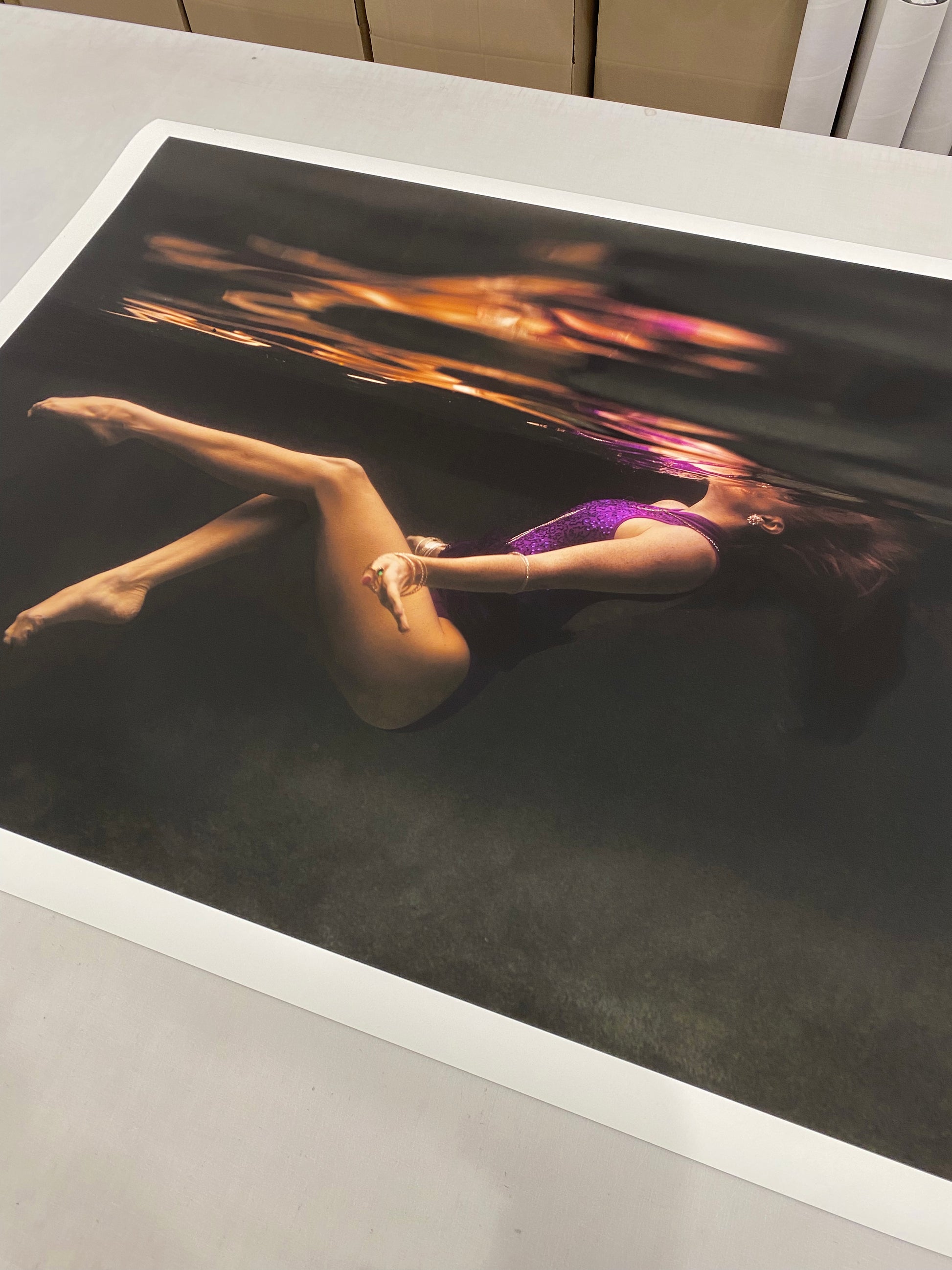 A large photograph from the "Show Time #2 Part 3" series by Francesca Owen, hand-signed by the artist, depicts a person floating on their back underwater in purple attire. The image rests on a table in a room with rolls of paper and cardboard boxes stacked in the background. The reflection of the person shimmers on the water's surface.