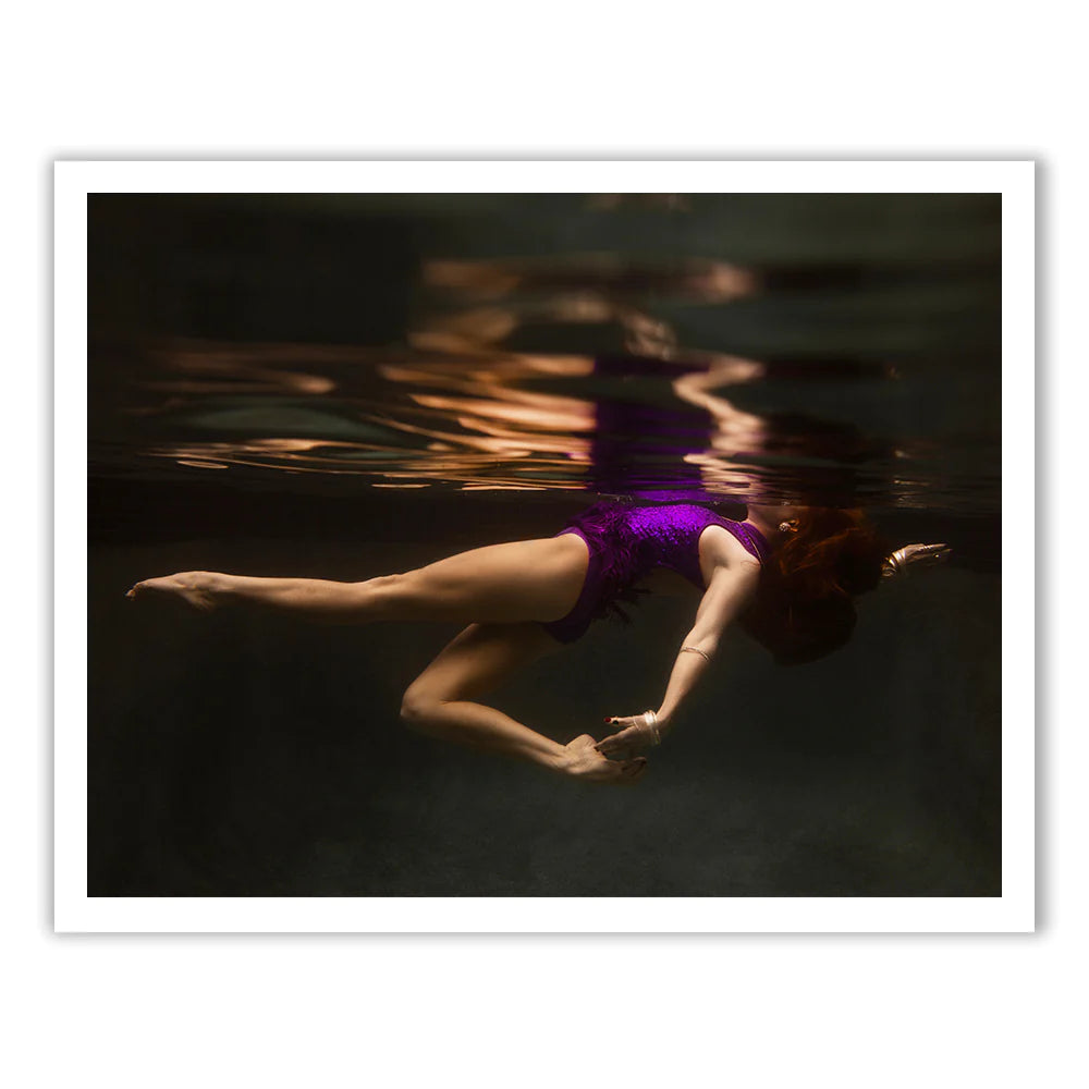 A woman in a purple swimsuit is floating underwater in a serene, dimly lit environment. Her body is gracefully curved, with one arm stretched forward and the other bent, creating a tranquil and weightless appearance. The scene's ethereal beauty would be perfectly captured on Francesca Owen's Show Time #2 Part 2 using fine art paper and archival pigments.