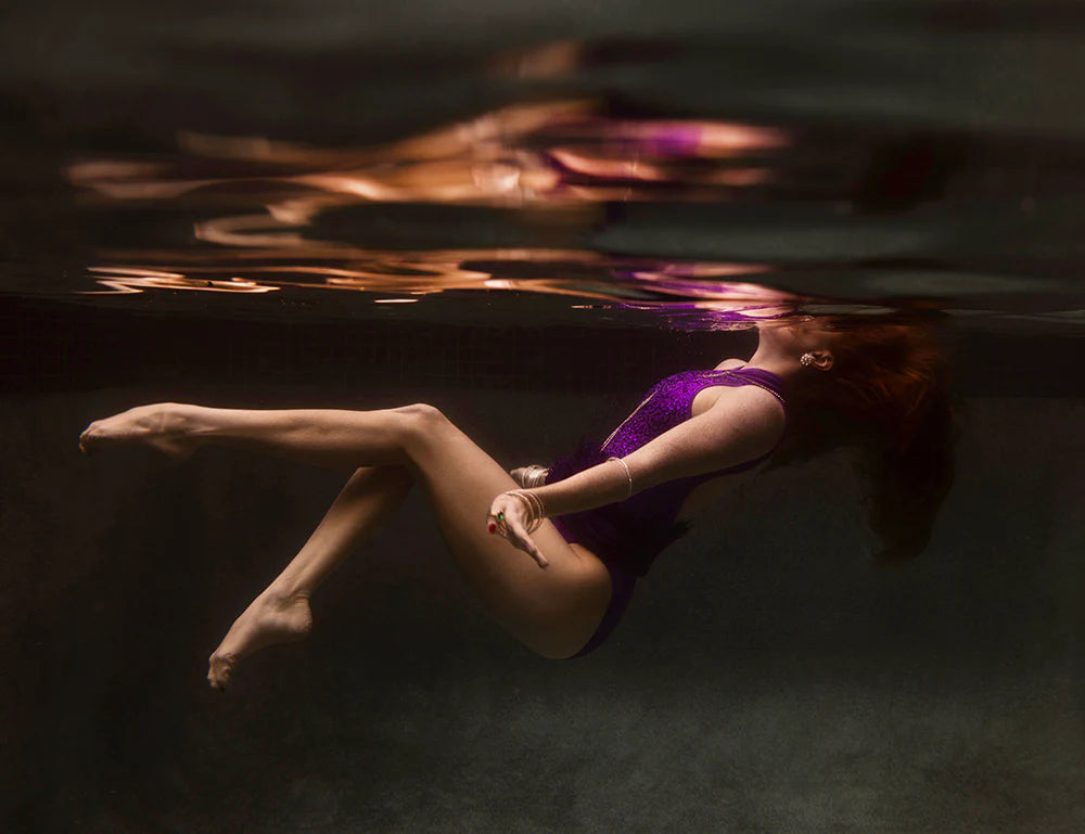 Underwater view of a person in a purple swimsuit floating effortlessly while upside down. Their red hair streams around them as they remain suspended in the water. The surface of the water reflects their image, creating a serene, ethereal scene captured in "Show Time #2 Part 3," an artwork by Francesca Owen, printed with archival pigments on fine art paper.