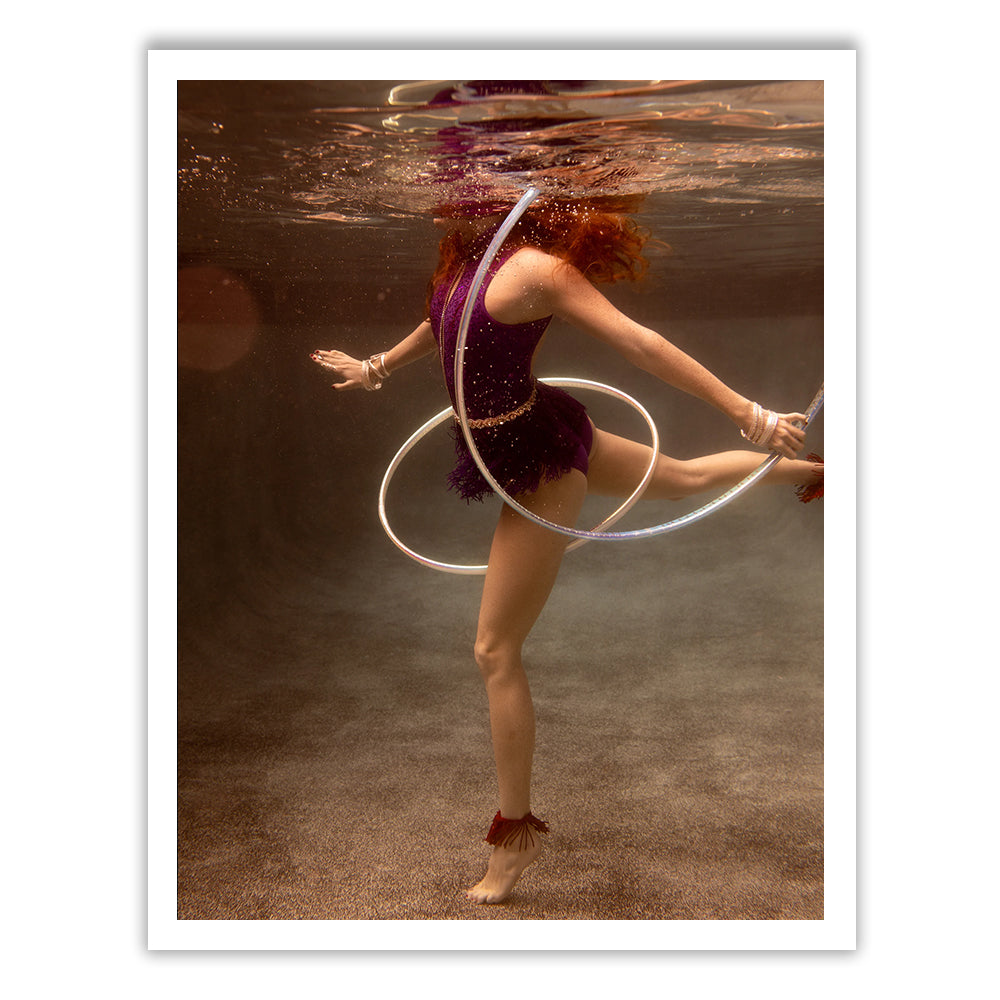 In "Show Time #5" by Francesca Owen, a red-haired individual in a purple outfit gracefully dances underwater, balancing on one leg while twirling a hula hoop. The slightly rippled water surface above them and the visible bottom floor are beautifully captured with archival pigments on fine art paper. This piece is available in custom sizes.