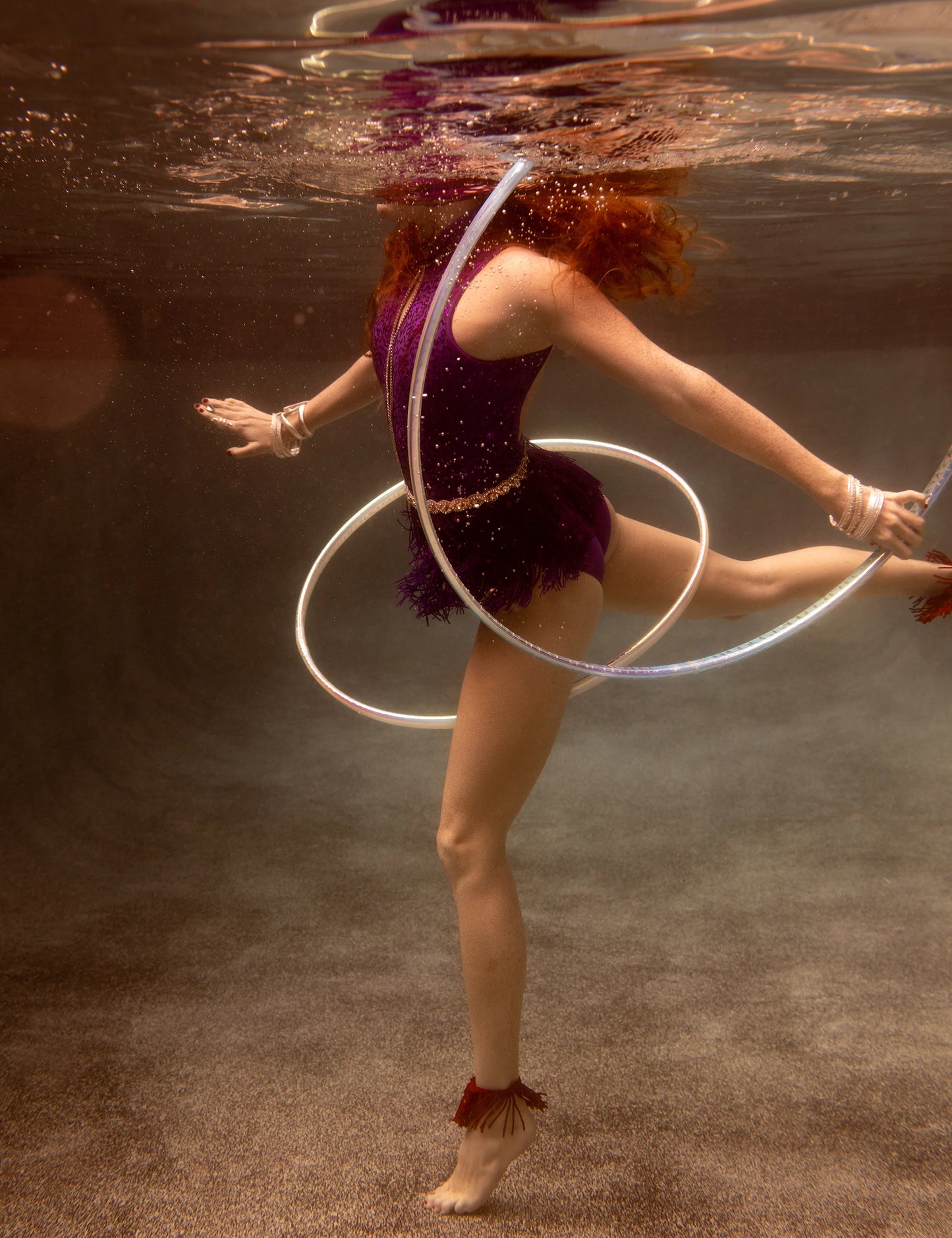 A person donned in a purple outfit gracefully performs an underwater dance, skillfully twirling a hula hoop around their waist. The serene scene is beautifully captured on fine art paper under soft illumination, showcasing the elegance of the performance. The person's red hair flows freely in the water, adding to the ethereal quality of "Show Time #5" by Francesca Owen.