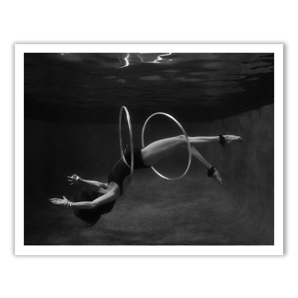 In Francesca Owen's "Show Time #9," a black and white underwater photo captures a person in a swimsuit gracefully arching their back, with two large hoops encircling their body, arms extended, and legs bent at the knees. Subtle ripples in the water above are beautifully rendered on fine art paper using archival pigments.