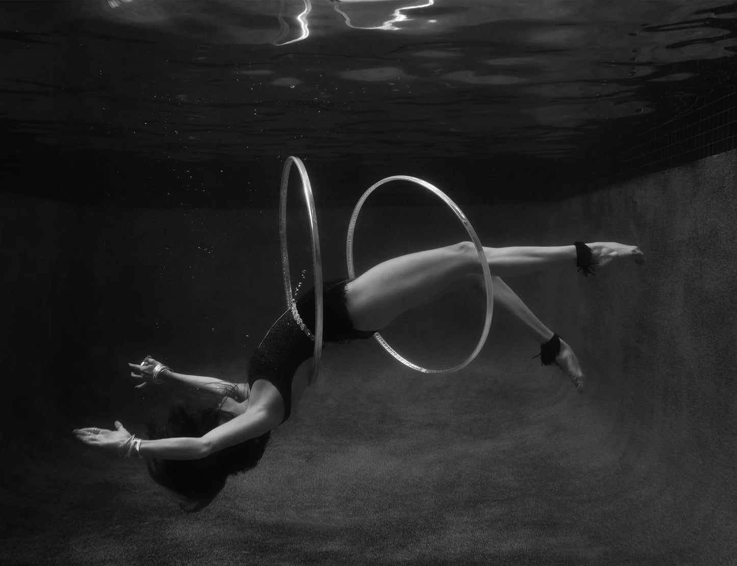 A black and white photo titled "Show Time #9" by Francesca Owen captures a person gracefully dancing underwater with two hula hoops. The individual is floating horizontally, with the hula hoops encircling their torso and legs, creating an ethereal scene in the dimly lit pool. A stunning example of fine art photography from Francesca Owen’s collection.