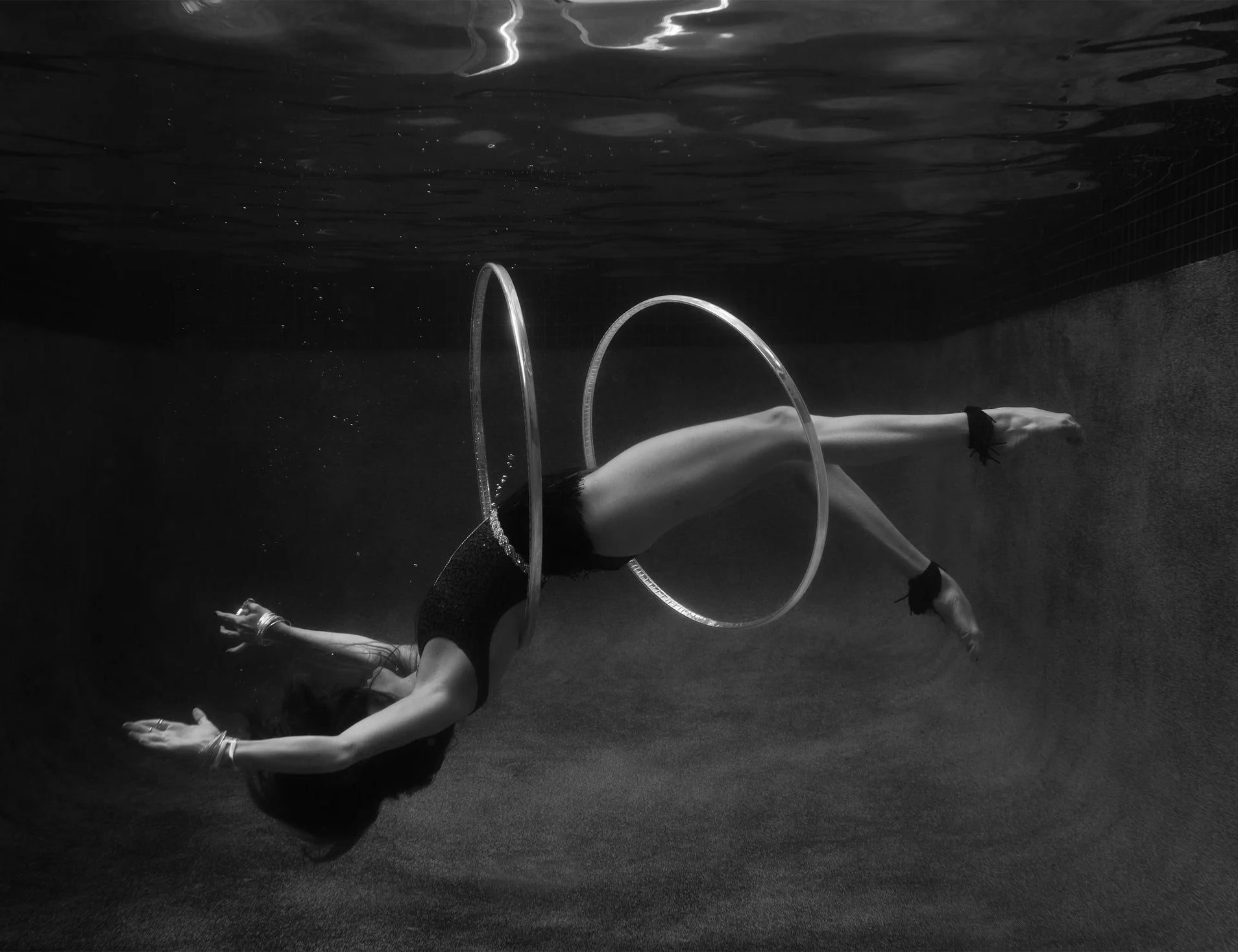 A black and white photo titled "Show Time #9" by Francesca Owen captures a person gracefully dancing underwater with two hula hoops. The individual is floating horizontally, with the hula hoops encircling their torso and legs, creating an ethereal scene in the dimly lit pool. A stunning example of fine art photography from Francesca Owen’s collection.