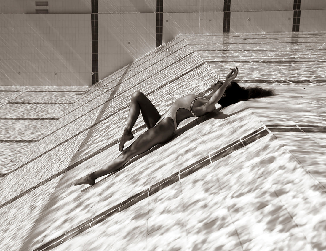 A person in a swimsuit lies on their back at the shallow end of an empty swimming pool. The black-and-white image, entitled "Flying Solo 03" by Francesca Owen, captures sunlight casting rippled water reflections on the tiles and the person's body. The artwork is printed with archival pigments and available in custom sizes, complete with a certificate of authenticity.