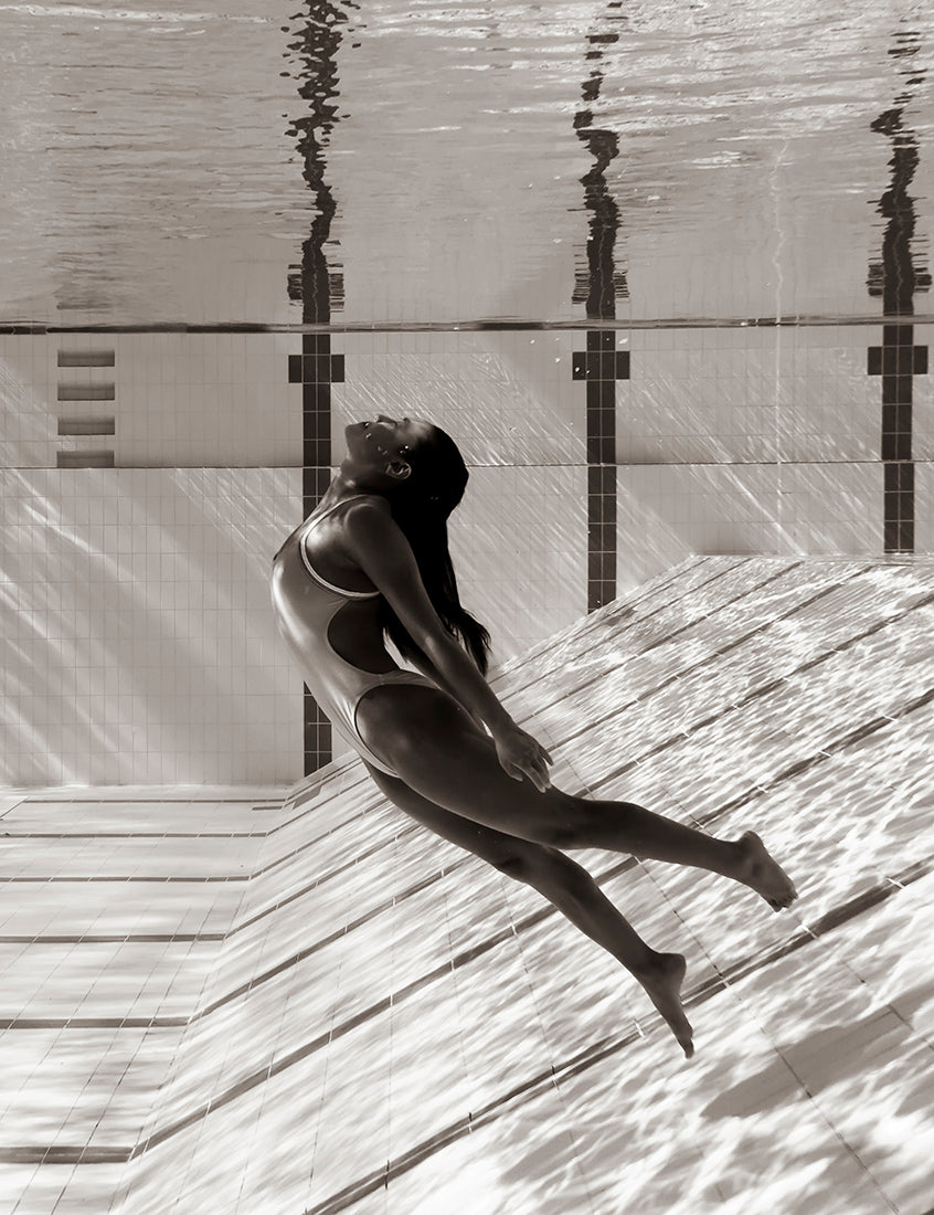 A woman in a one-piece swimsuit gracefully floats underwater with her back arched and arms stretched alongside her body. The water ripples create reflections on the pool tiles, enhancing the serene underwater scene. "Flying Solo 06" by Francesca Owen, printed on fine art paper with archival pigments, is truly striking.