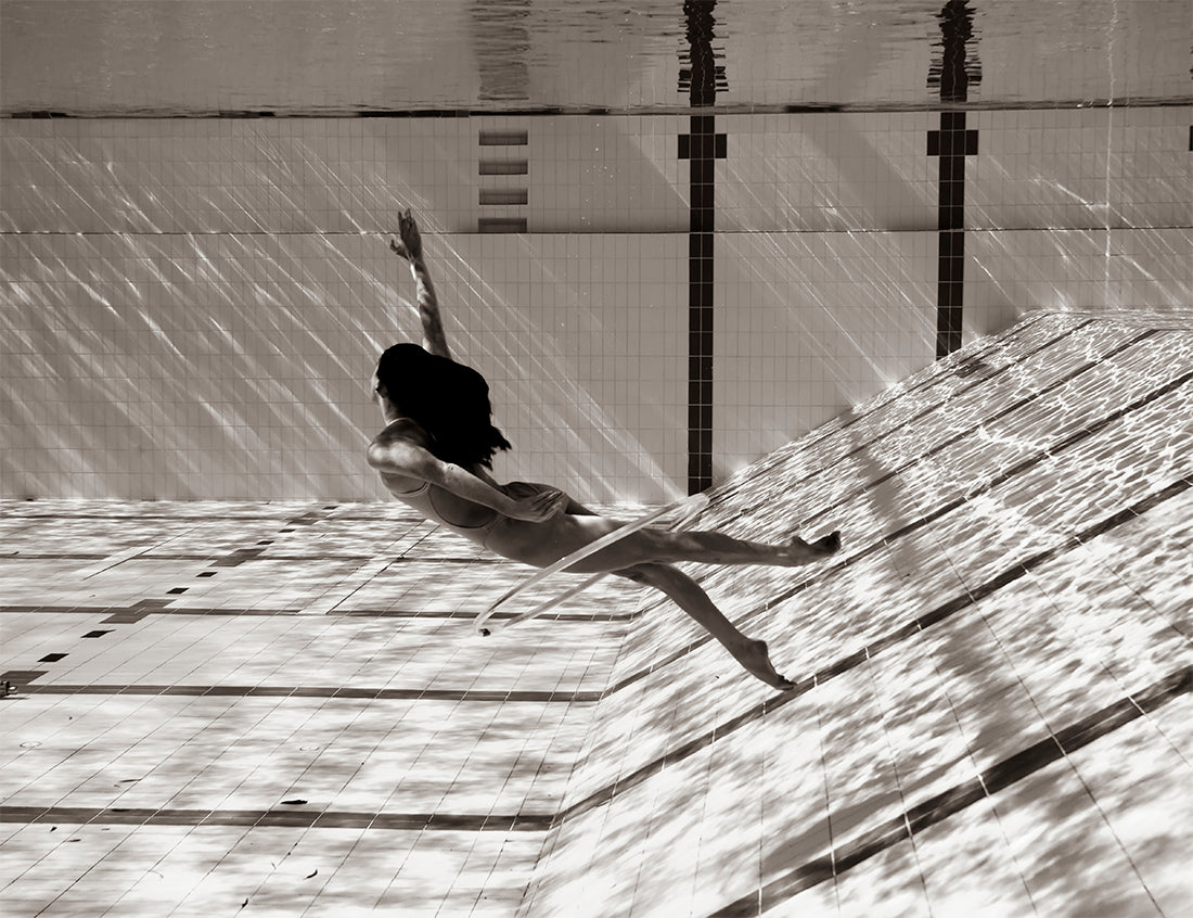 A woman swims underwater in a tiled pool, gracefully reaching upward. The water's surface reflects light patterns onto her silhouette, creating a mesmerizing play of light and shadow. "Flying Solo 08" by Francesca Owen is available as fine art prints with archival pigments, each piece accompanied by a certificate of authenticity.