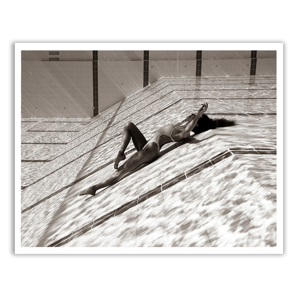 A black and white image from the "Flying Solo 03" collection by Francesca Owen features a woman lying on her back in a swimming pool. Her body creates a diagonal line within the pool's geometric tiles, with shimmering water effects on her body and surrounding surfaces, captured using archival pigments to ensure longevity.