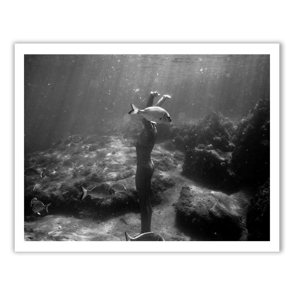 Still Waters #13 by Francesca Owen captures a black and white underwater scene. It depicts a person submerged, standing upright with arms raised towards the surface, surrounded by various fish swimming nearby. The background features rocky formations illuminated by sunlight filtering through the water. Available as a fine art print with a certificate of authenticity.