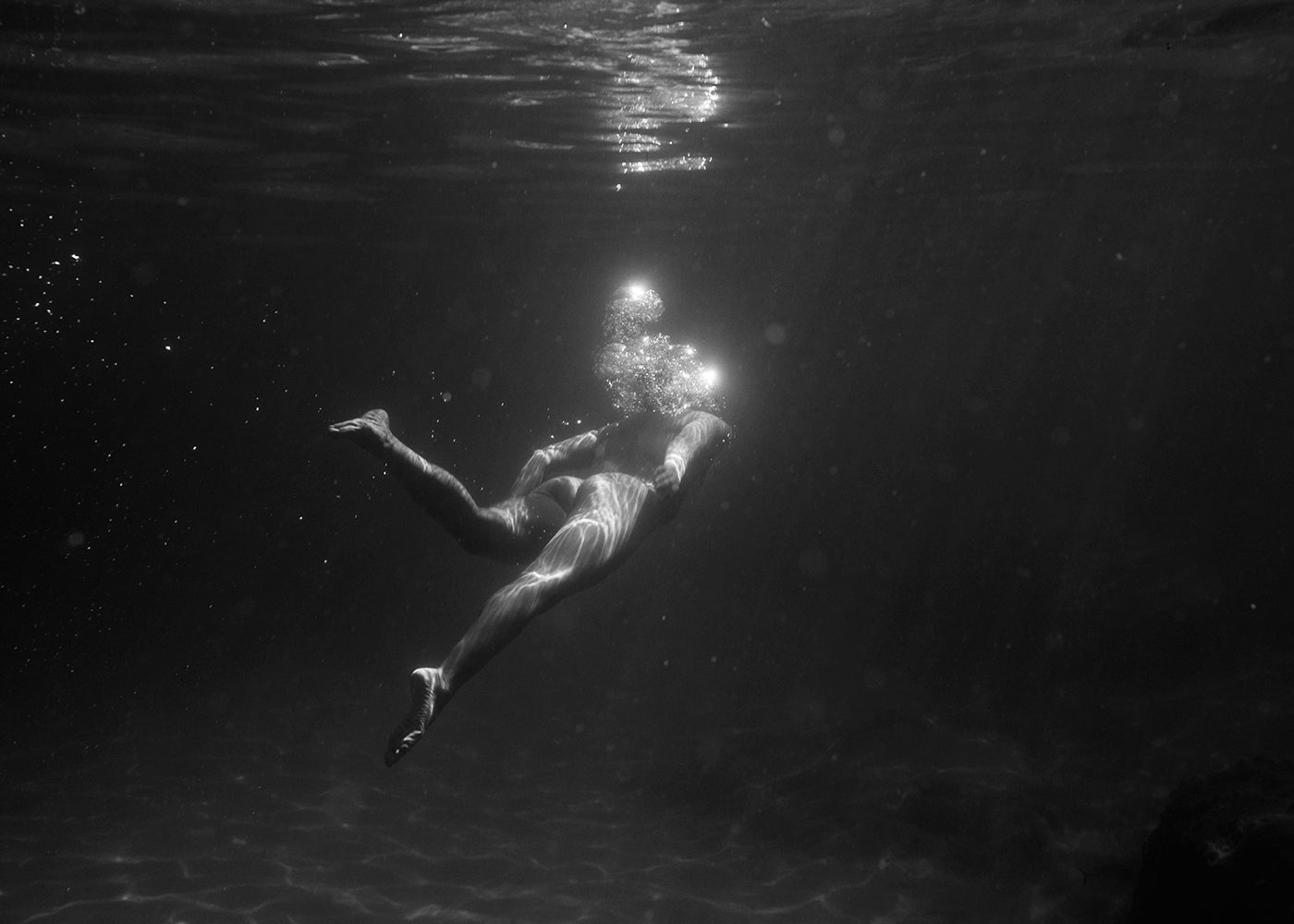 Still Waters #4 by Francesca Owen is a black and white fine art print depicting a person swimming underwater with sunlight filtering from above. The image, printed on fine art paper using archival pigments, captures the swimmer moving horizontally, creating bubbles around their head and light reflections on their body in a dark, mysterious underwater environment.