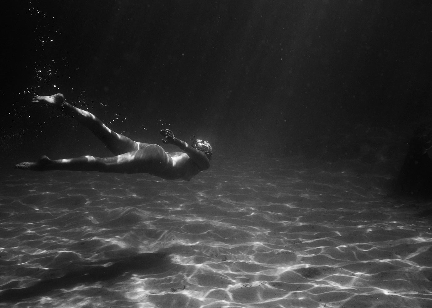 Experience the captivating elegance of Still Waters #7 by Francesca Owen. This black and white photograph beautifully captures a person underwater with sunlight filtering down from the surface, illuminating the swimmer and casting intricate patterns on the sandy bottom. The swimmer is depicted in motion, caught mid-swim with bubbles trailing behind. Perfect for custom framing as fine art prints using archival pigments.