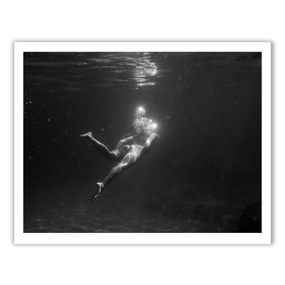 Still Waters #4 by Francesca Owen is a black and white photo depicting a person swimming underwater. Sunlight filters through the surface, illuminating the serene swimmer as they float in the dark water. Printed on fine art paper with archival pigments, this image beautifully captures a sense of calm and tranquility. The photograph includes a certificate of authenticity.