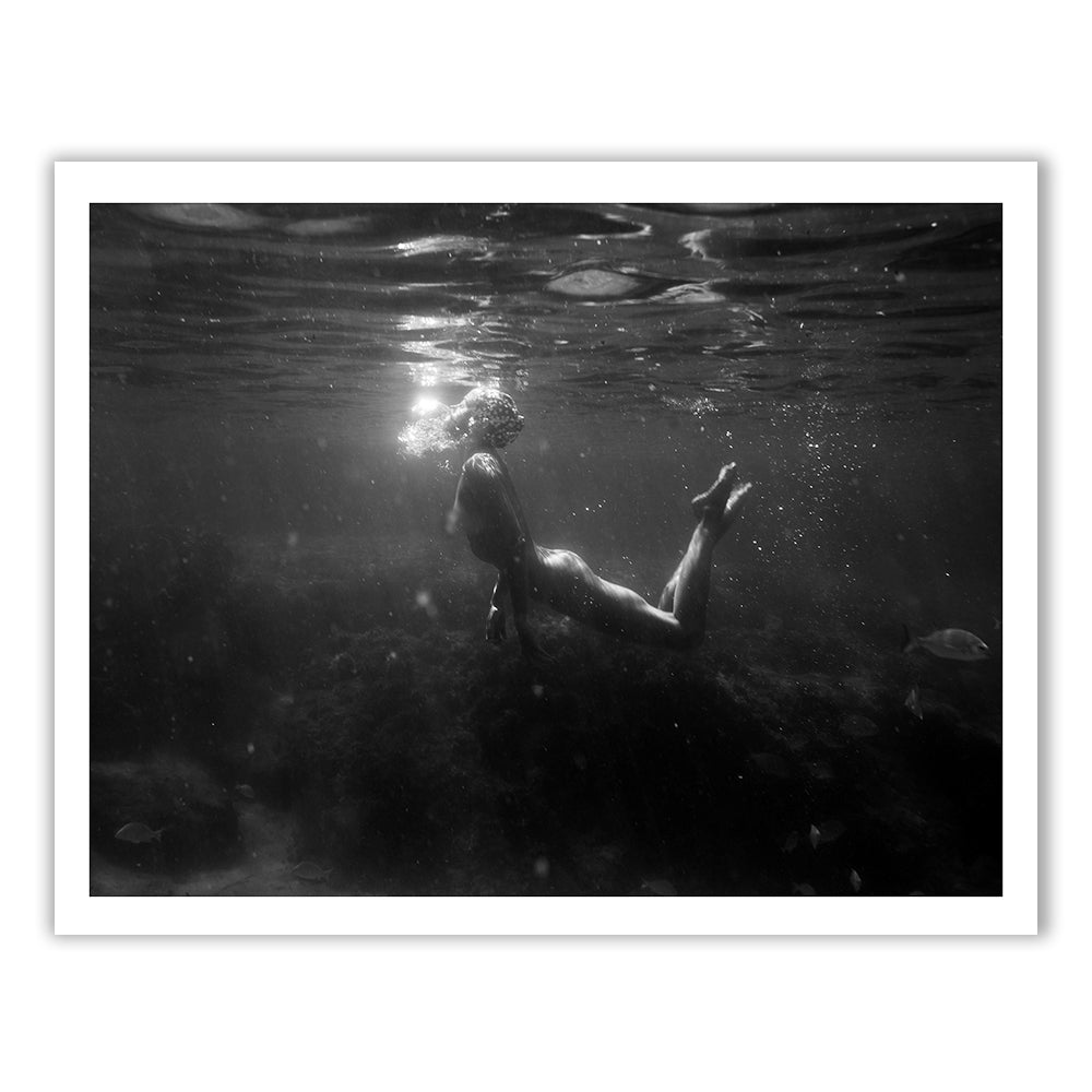 Still Waters #11," a captivating piece by Francesca Owen, portrays a black and white photograph of a person swimming underwater and facing upwards in a murky, dimly lit environment. Partially illuminated, the swimmer exhales bubbles towards the surface while small fish are faintly visible in the background. This mesmerizing image is available as fine art prints with worldwide shipping.