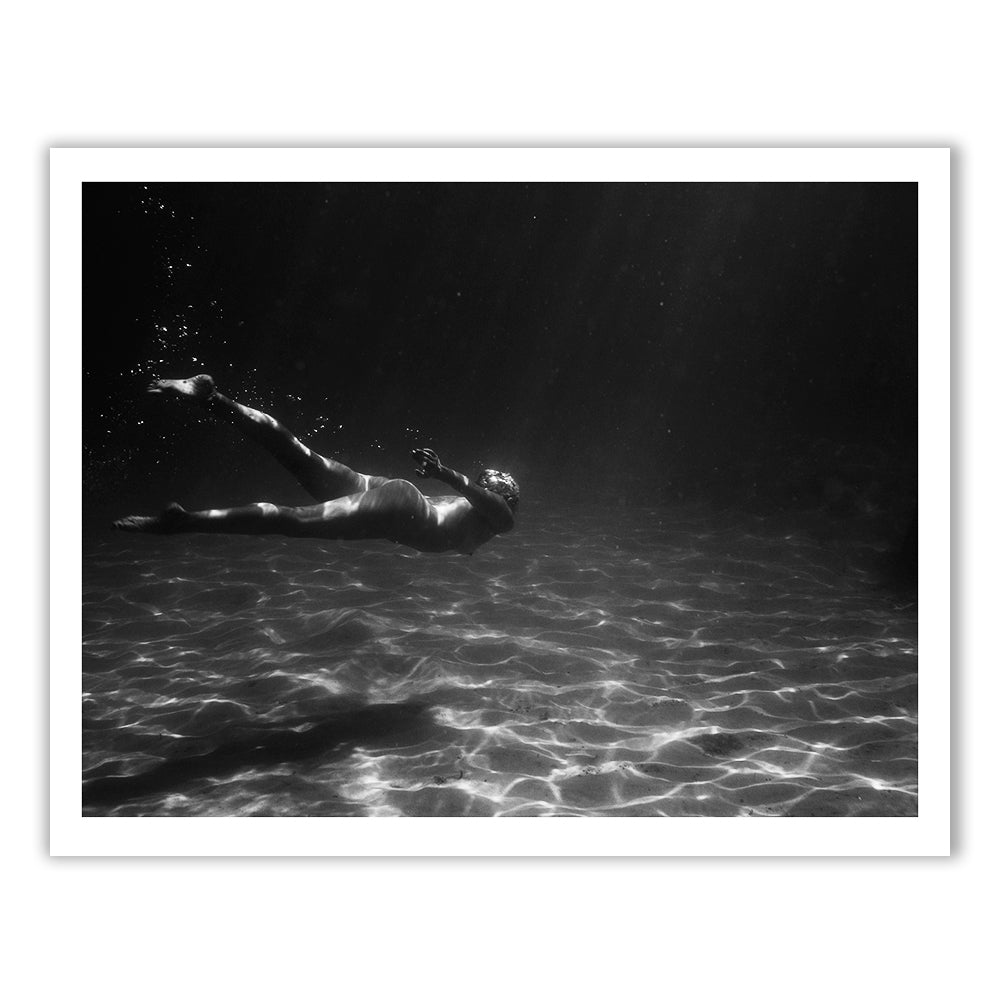 Still Waters #7 by Francesca Owen features a black-and-white image of a person swimming underwater in an ocean or pool. Sunlight penetrates the water, creating a dappled pattern on the sandy bottom. The swimmer is moving horizontally, with bubbles trailing from their movements. This captivating scene is perfect for fine art prints and custom framing using archival pigments.