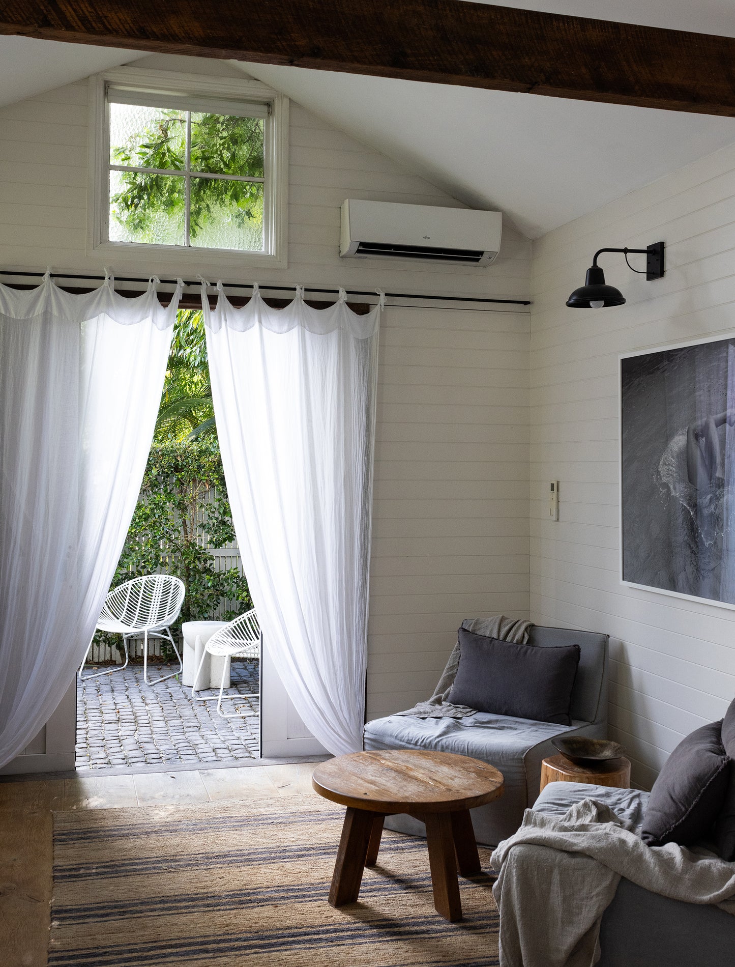 A cozy living room with a wooden beam ceiling features a custom-made piece from the Francesca Owen collection, called "Self Reflection," which serves as a charming small round table. Cushioned chairs provide comfortable seating, while white sheer curtains partially cover a glass door that opens to a patio adorned with wicker chairs and lush greenery. Above the door, an air conditioner ensures comfort throughout the space.