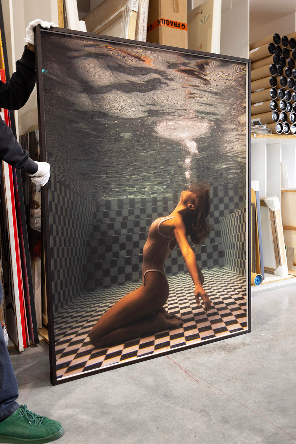 A person wearing white gloves holds a large framed photograph titled *The Banya #1* by Francesca Owen. It depicts a woman in a one-piece swimsuit posing underwater in a pool with a checkered pattern on the floor and walls. The woman is exhaling bubbles while arching her back. This fine art print boasts archival pigments for longevity and custom framing for elegance.