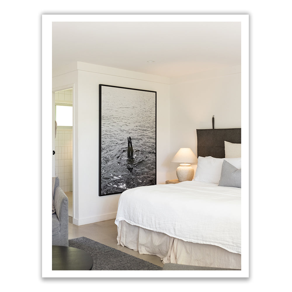 A modern bedroom showcases the Capri Tippy Toes, a large bed adorned with white linens from Francesca Owen, alongside a matching bedside table and white lamp. A striking framed photograph on the wall is printed on fine art paper using archival pigments. The room's soft, neutral tones offer a hint of an adjacent tiled bathroom.