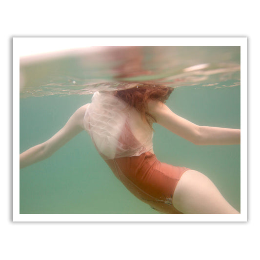 A person swims underwater in a reddish-orange swimsuit, with a translucent white fabric draped over their shoulder. The clear water and the soft, ethereal quality of the image are reminiscent of Francesca Owen's "Softness #1 B&W (Copy).