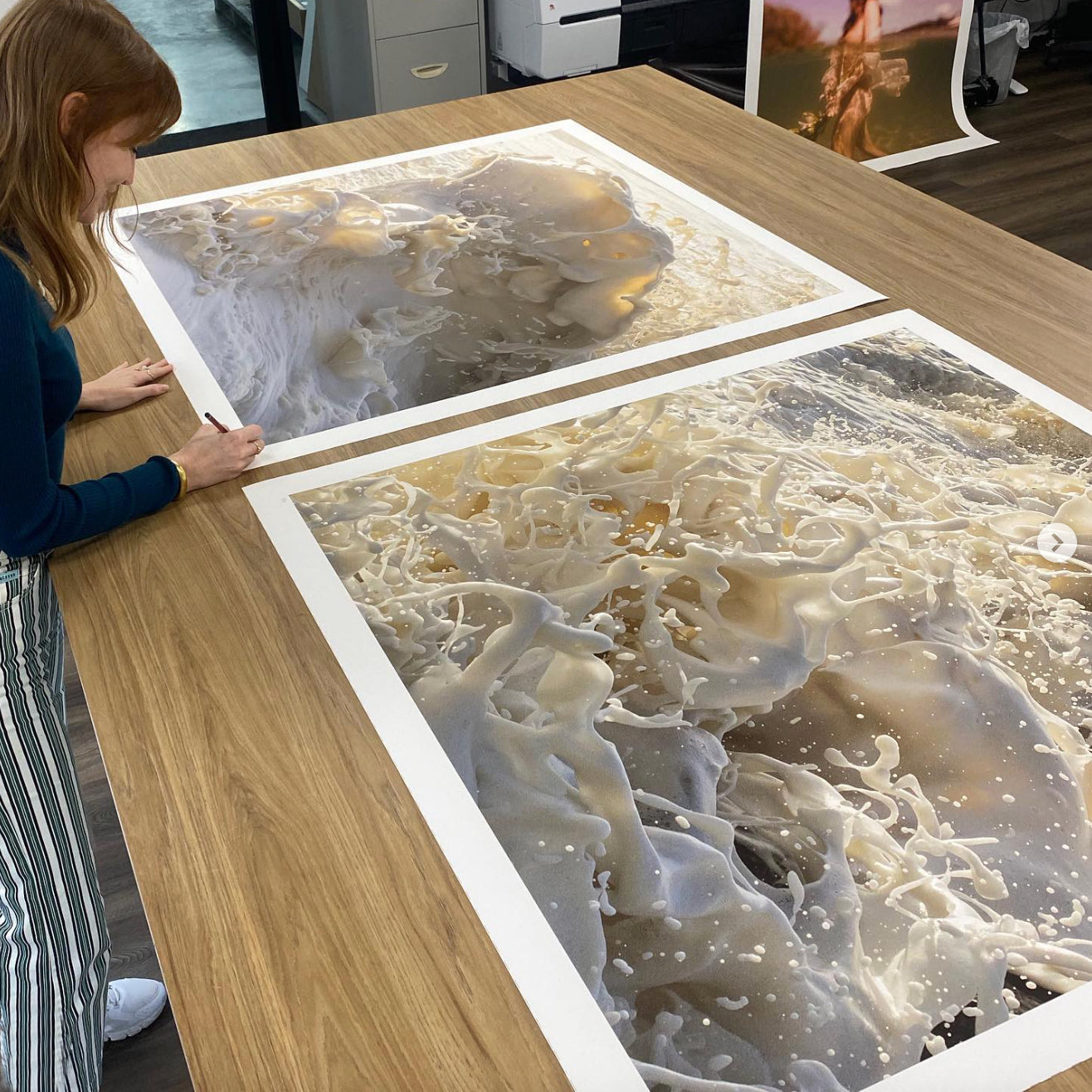 A person with long hair and a dark long-sleeve shirt examines two large, vibrant prints titled "Wild Seas Part I, V 2 — Limited Edition Print" by Francesca Owen, laid out on a wooden table. The intricate details and textures of these archival pigment prints on fine art paper are striking. Additional equipment and a framed picture can be seen in the background.