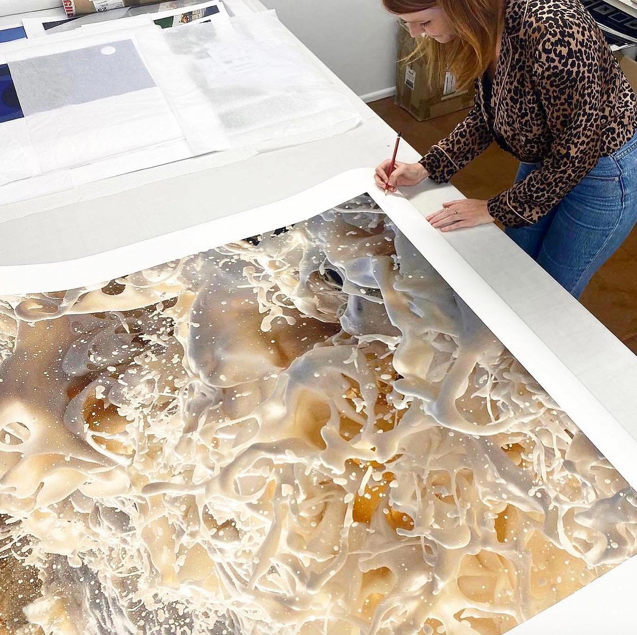 A person in a leopard print top is using a pencil to draw on a large, abstract artwork laid out on a table. The artwork, destined to become the "Wild Seas Part I, V 2" limited edition print by Francesca Owen, features swirling patterns in beige, white, and brown tones. Various other art pieces and materials are visible in the background.