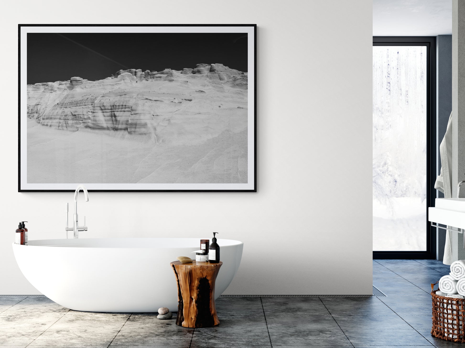 A modern bathroom featuring a freestanding white tub, complemented by a wooden stump side table adorned with bath products, and "Magic in Milos #14," a large framed black-and-white landscape photo on fine art archival paper by Francesca Owen. A window reveals a snowy scene outside.