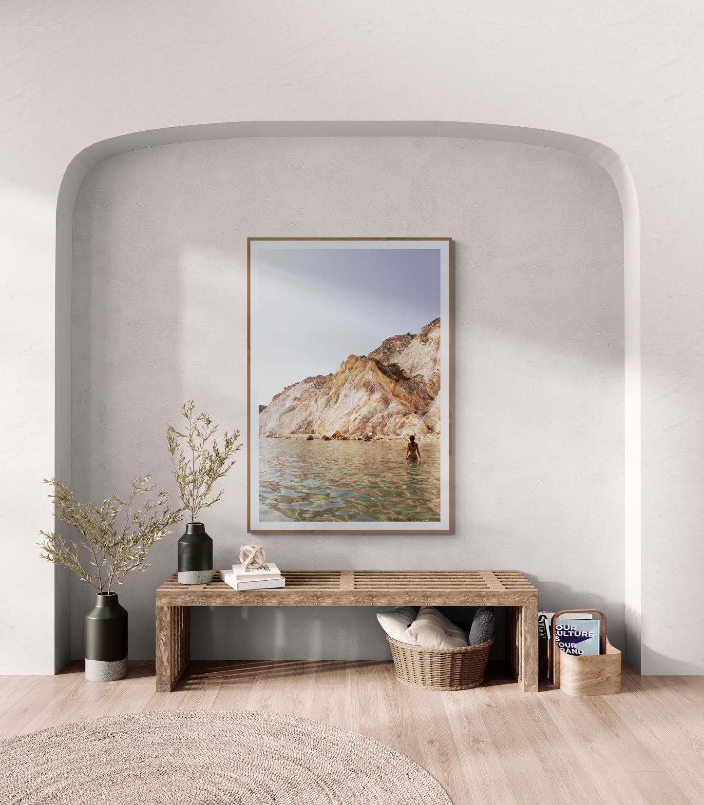 A minimalist room showcases Francesca Owen's "Magic in Milos #11," a framed landscape photo depicting a rocky coast, printed on fine art paper with archival pigments, placed above a sleek wooden bench. The bench is embellished with a small vase of flowers, two pillows, and a wicker basket. Neutral tones and gentle lighting contribute to the peaceful ambiance.