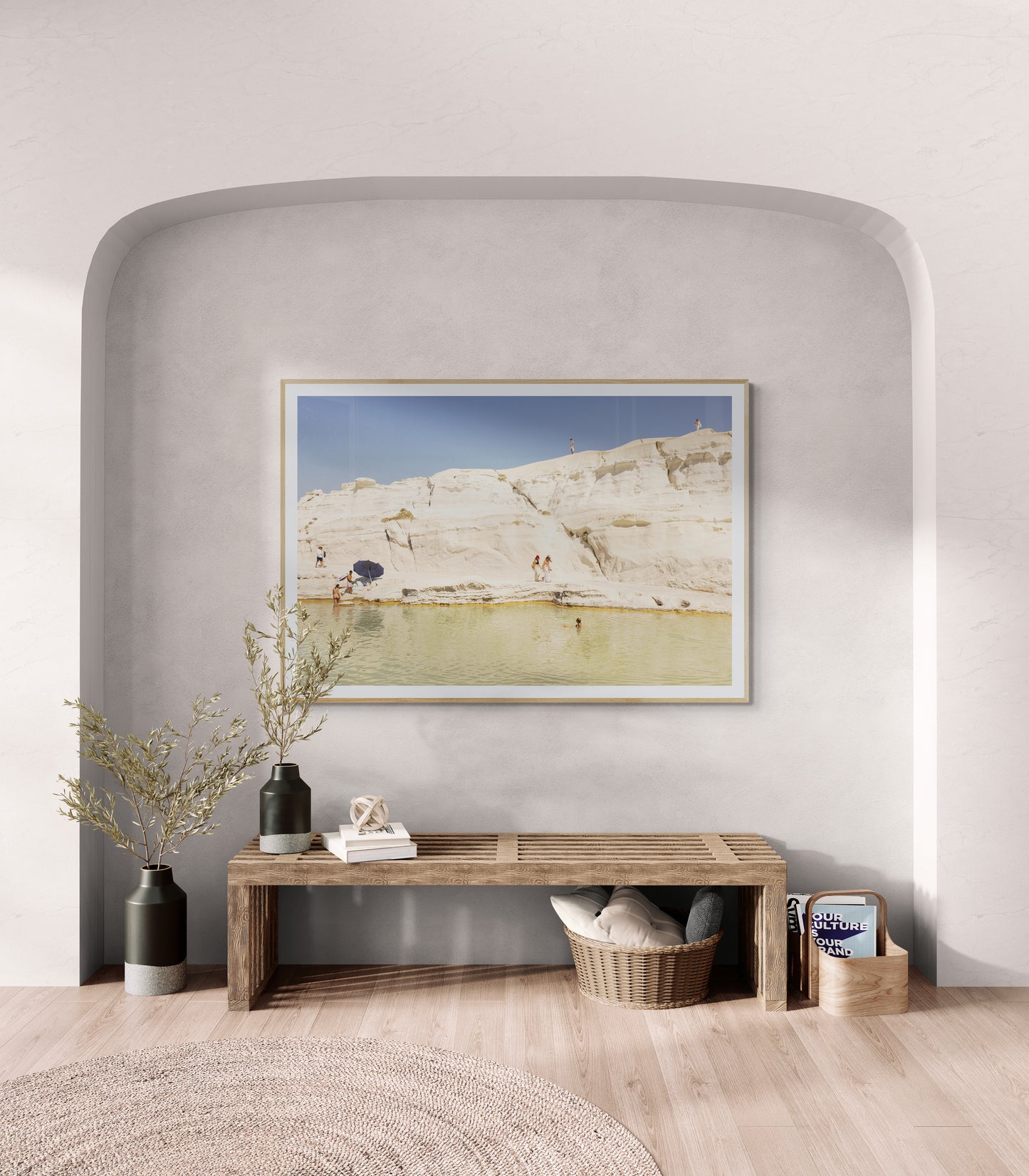 A minimalist room showcases a wooden bench made from Tasmanian Oak placed beneath a framed painting titled "Magic in Milos #13" by Francesca Owen, which captures a beige rocky landscape with water on archival rag paper. The bench is adorned with two dark vases and a basket filled with books, while light streaming in from the left adds warmth and invitation to the space.