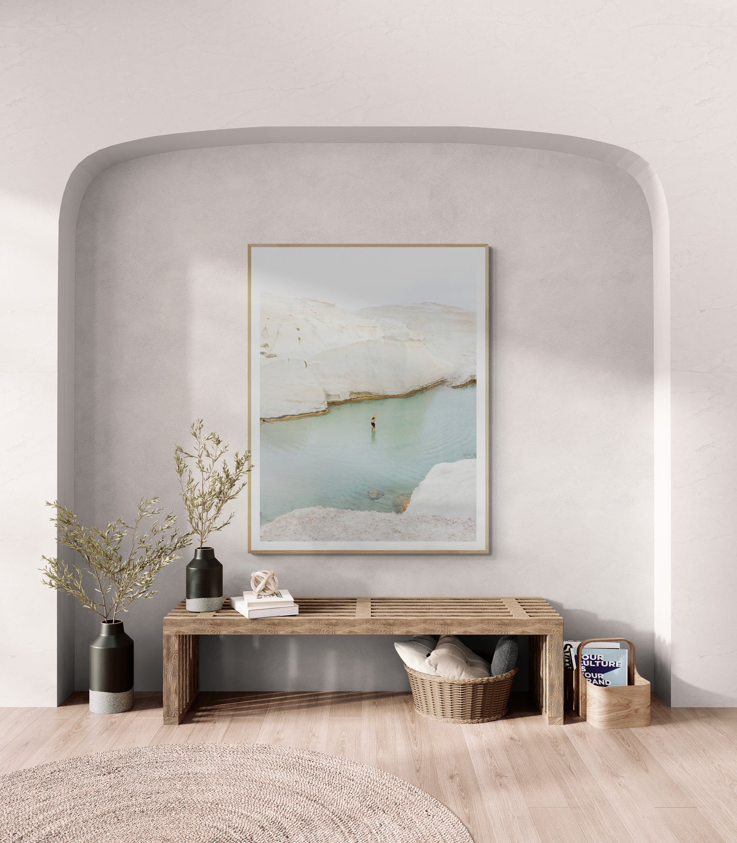 A cozy room features "Magic in Milos #2," a large framed photo by Francesca Owen, showcasing a serene coastal scene printed on fine art paper with archival pigments, gracing an arched wall. Below, a wooden bench holds a vase with branches, a candle, and a basket. A round rug and natural light enhance the tranquil atmosphere.