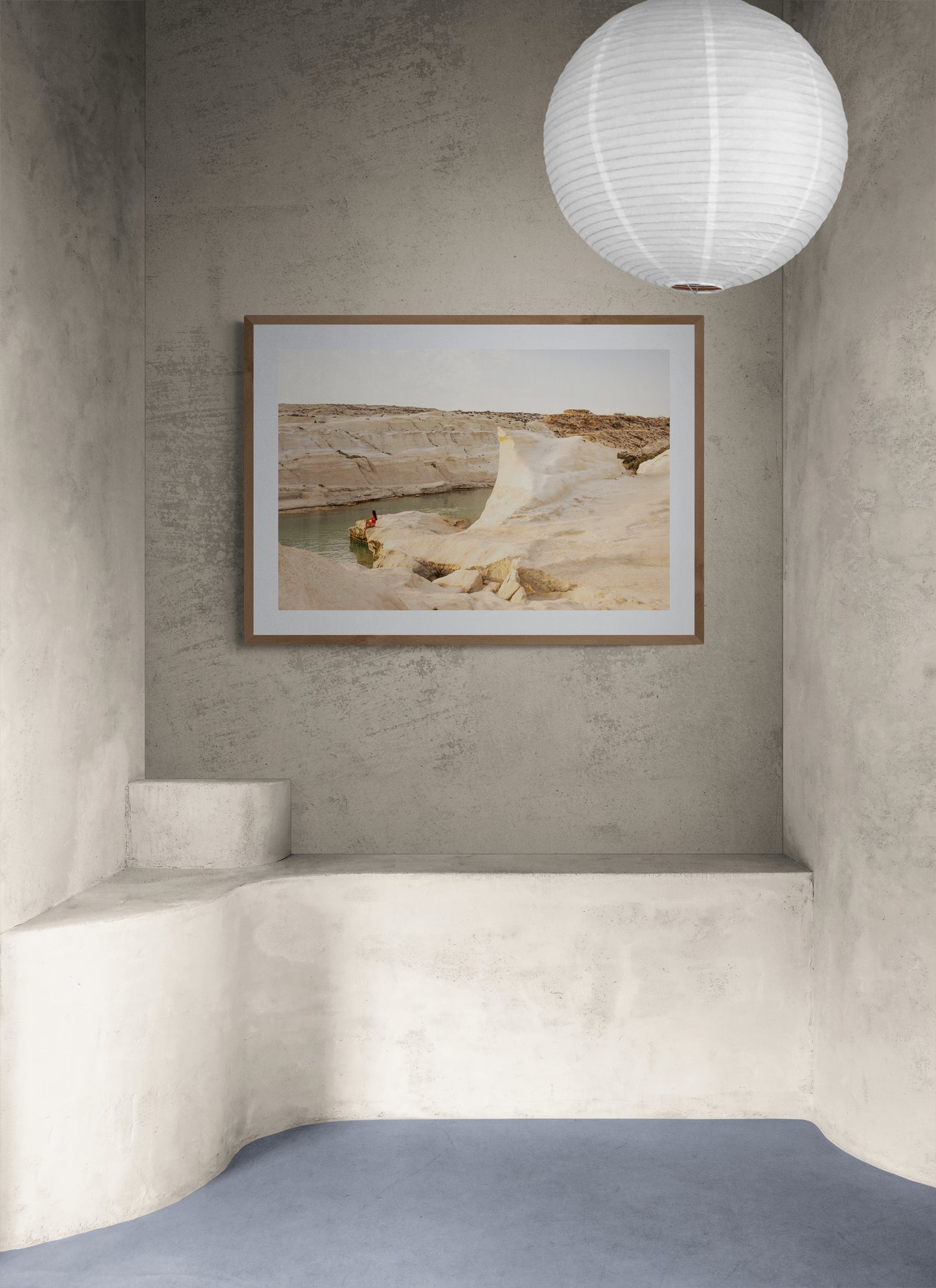 A minimalist room with textured grey walls and a smooth floor showcases "Magic in Milos #5," a framed landscape photograph by Francesca Owen, depicting a rocky desert and river printed on fine art paper. A white, round paper lantern hangs from the ceiling, and a built-in bench is visible along the corner.