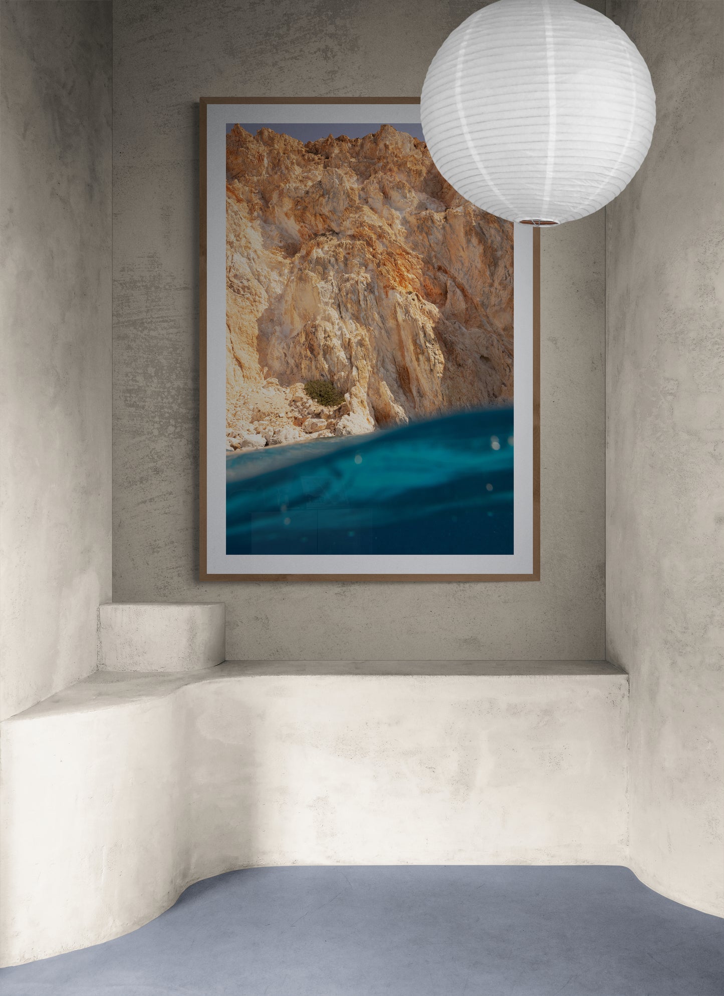 A serene room features "Magic in Milos #7," a large framed photo by Francesca Owen depicting a rocky cliff above turquoise water, printed on archival rag paper and adorning the textured gray wall. A white paper lantern hangs from the ceiling above a built-in concrete bench that curves along the walls, while the smooth floor is finished in muted blue tones.