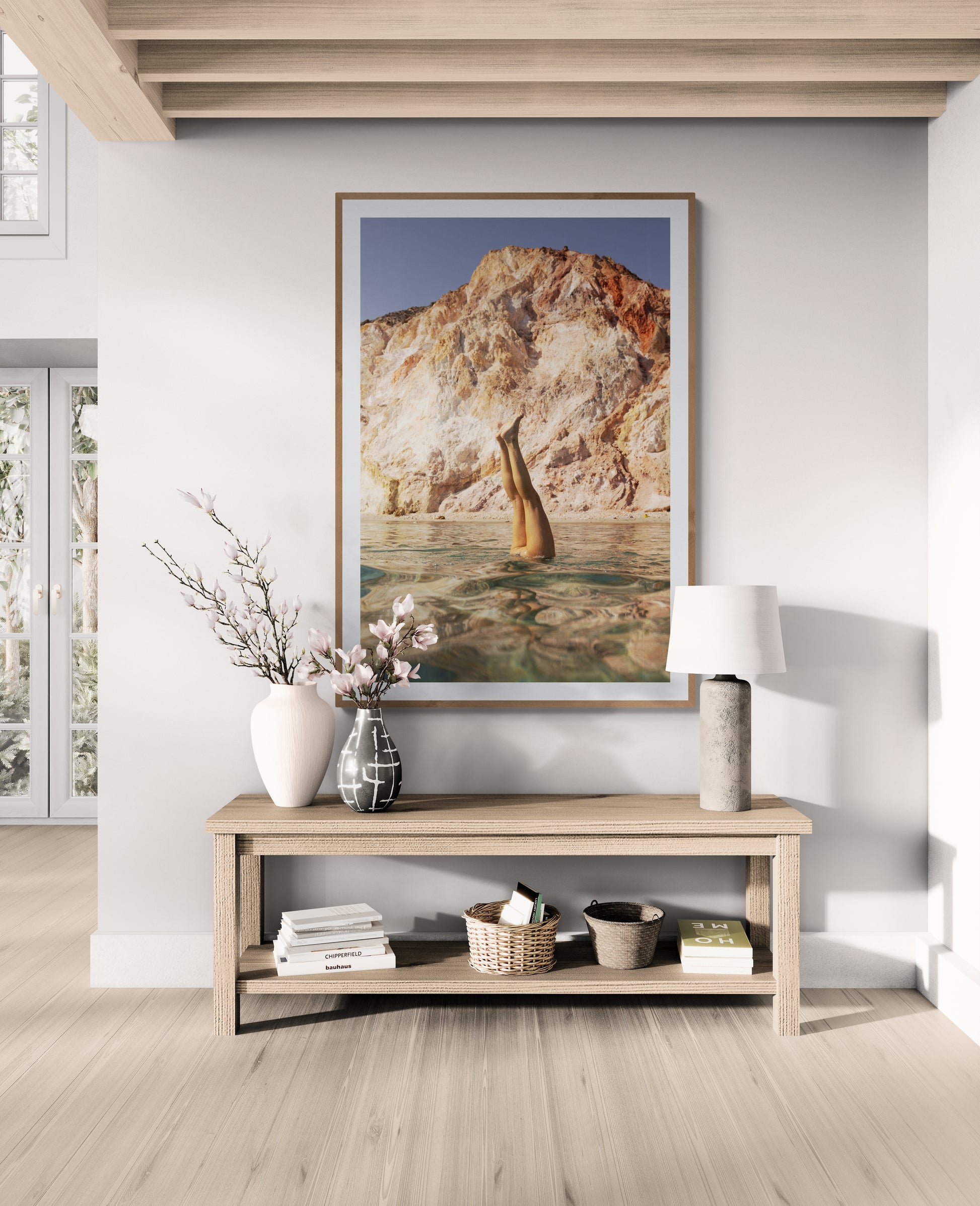 A bright, minimalist living room showcases a wooden console table decorated with a white vase of flowers and a lamp. Hanging above it is "Magic in Milos #10," a striking photograph by Francesca Owen, printed with archival pigments, featuring a person doing a handstand in the water against the backdrop of rocky cliffs.