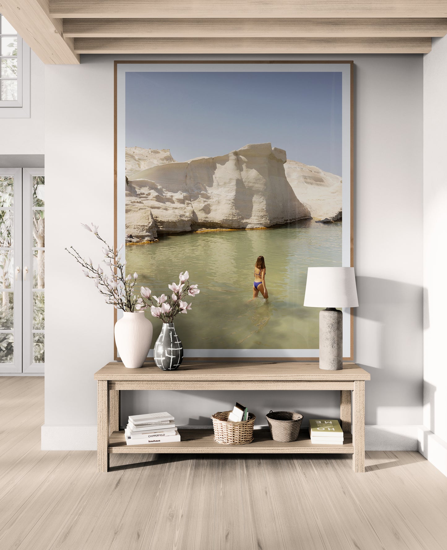 A modern living room features "Magic in Milos #12" by Francesca Owen, a large framed photograph in fine art archival print, illustrating a woman in green water amid white rocky cliffs. The artwork, framed in Tasmanian Oak, is displayed above a wooden bench adorned with a vase of flowers, books, and baskets. Natural light floods the room.
