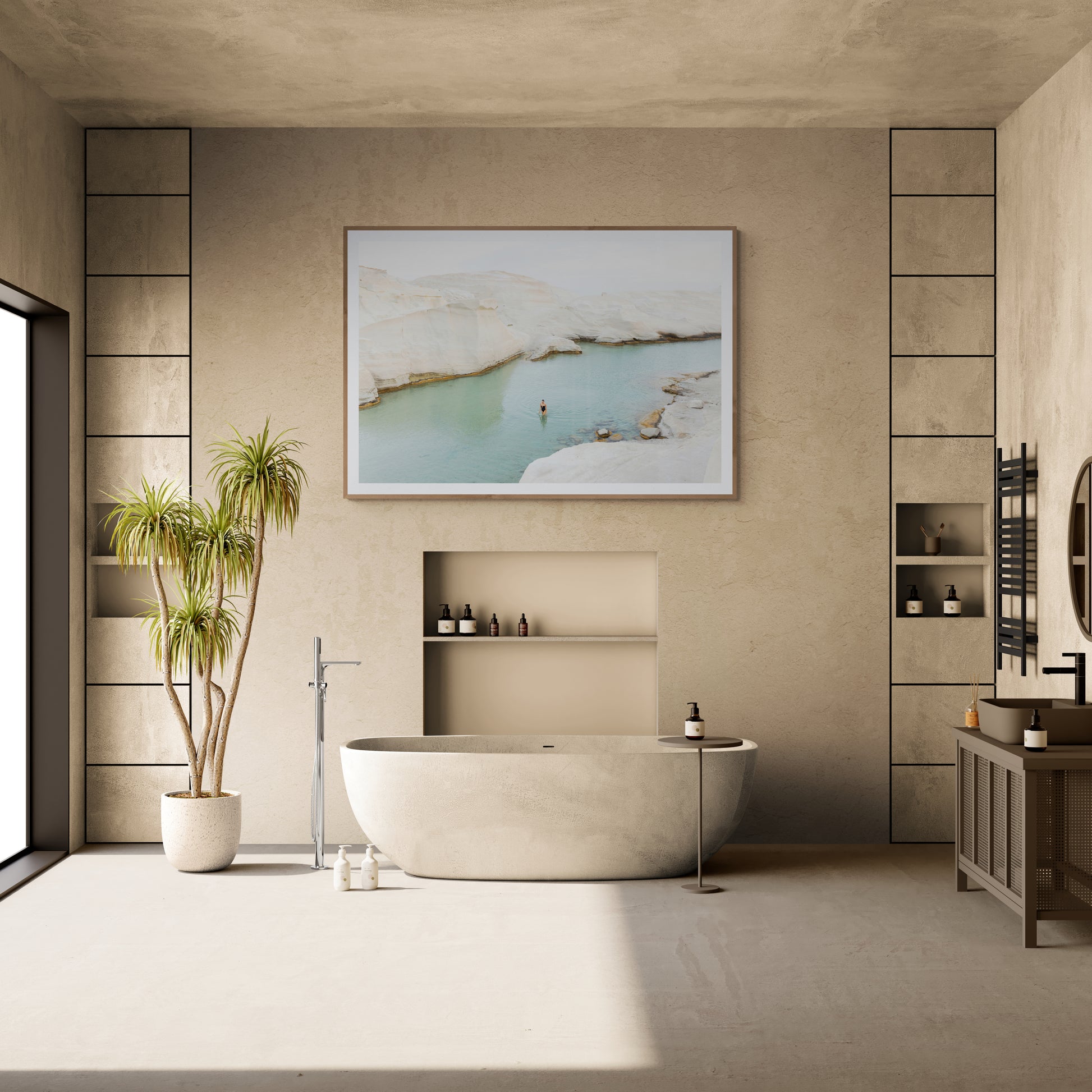 A contemporary bathroom is highlighted by the "Magic in Milos #1," a freestanding oval bathtub designed by Francesca Owen, featuring a captivating landscape photo printed on fine art paper to ensure enduring beauty. To the left, a tall potted plant complements the scene, while a vanity with a round mirror enhances elegance, all set against neutral tones and minimalist decor to create a serene ambiance.