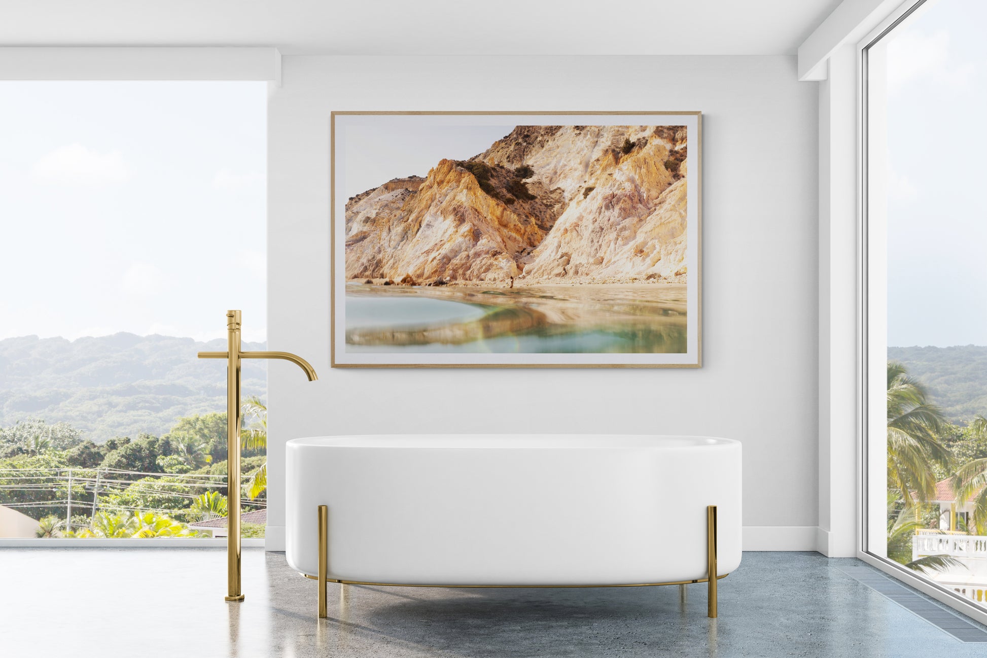 A modern bathroom features a freestanding white tub adorned with gold fixtures. A large window reveals a view of lush greenery, while "Magic in Milos #9," a fine art archival print by Francesca Owen with custom framing options, hangs above the tub, showcasing rocky hills and a calm water scene. The space is bright and minimalistic.