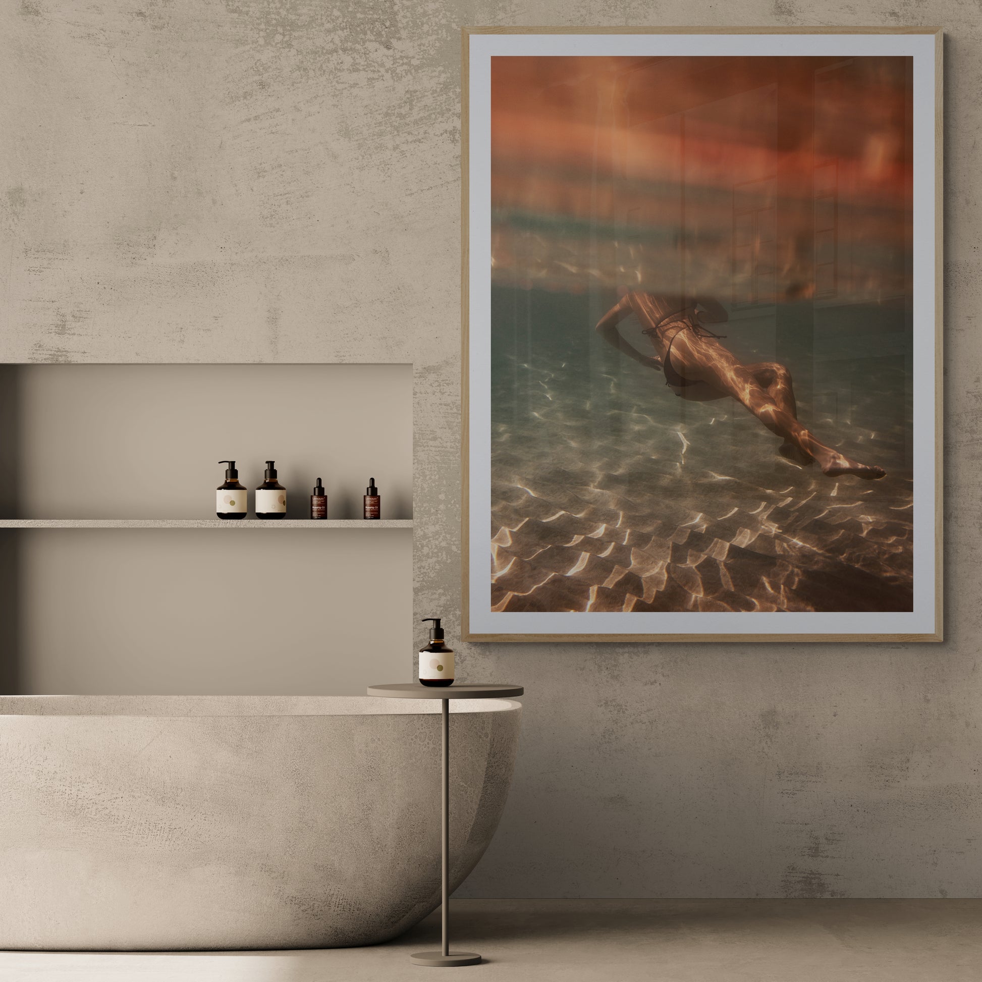 A minimalistic bathroom featuring a smooth stone bathtub, accompanied by a small round side table holding a soap dispenser and built-in shelves with bottles. The space is beautifully accented by "Magic in Milos #3" from Francesca Owen, a large framed photo printed on fine art paper showcasing a person swimming underwater, adding an artistic warmth to the atmosphere.