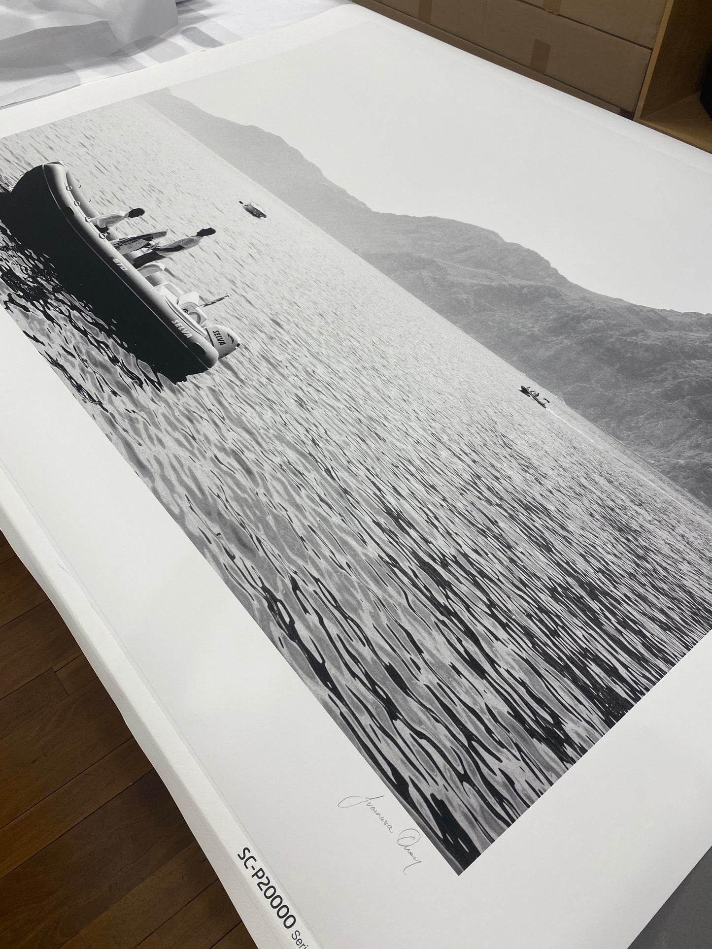 A large black and white photograph from the "Cruising" collection by Francesca Owen lies on a table. The serene lake scene, captured on fine art paper, shows people relaxing in a small boat, surrounded by distant mountains. Several other boats are scattered across the water. Hand signed with archival pigments, the signature and model number are at the bottom.