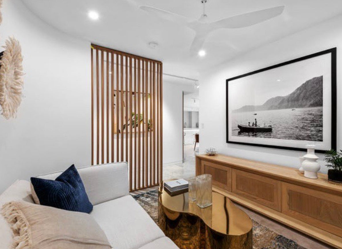 A modern living room with a white sofa, blue cushion, and brass coffee table. The room features "Cruising," a large framed black-and-white photo of a boat on archival pigments and fine art paper, hand-signed by artist Francesca Owen, displayed above a wooden console table. A wooden slat partition and ceiling fan enhance the contemporary decor.