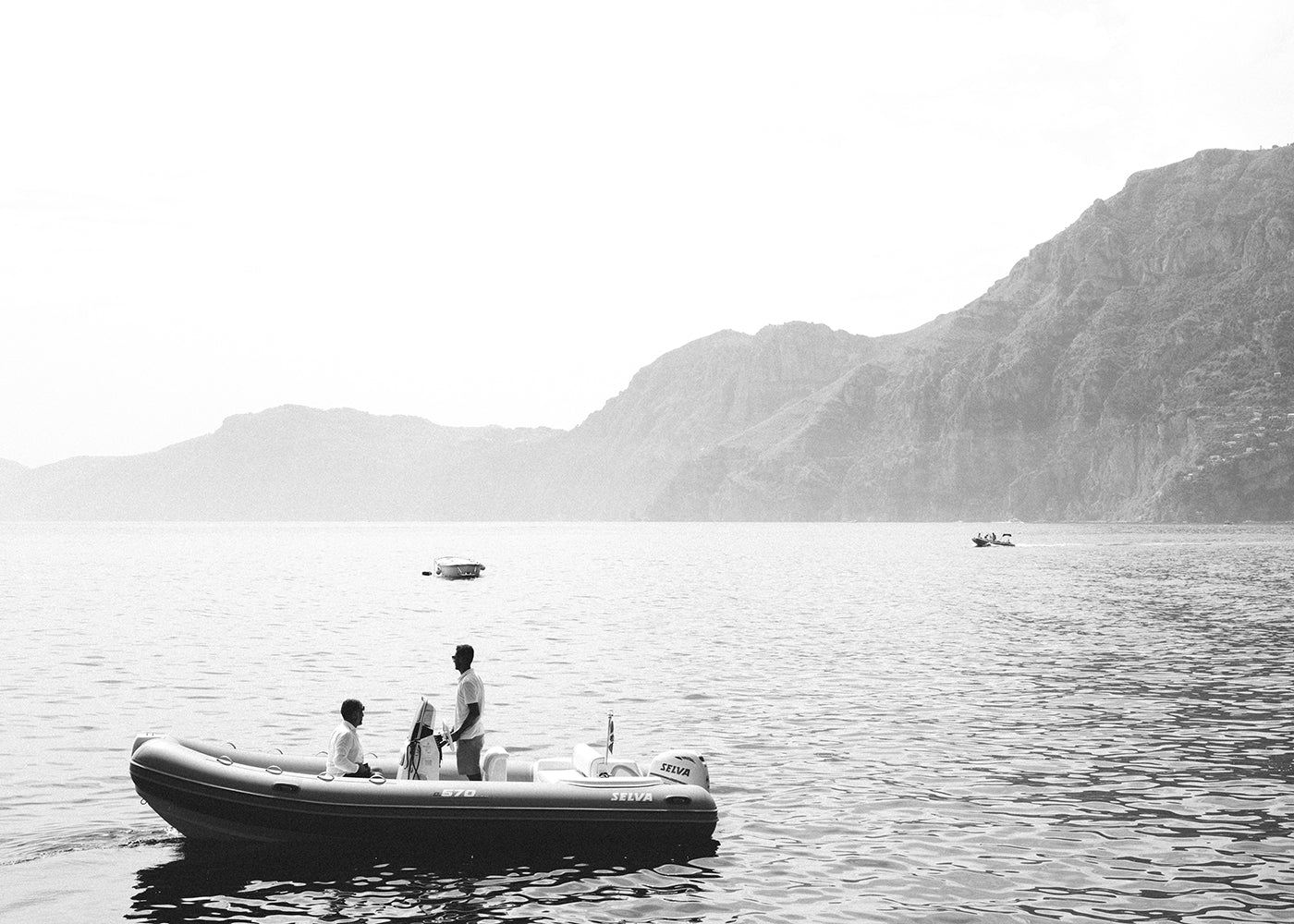 A black and white image of two people standing on a small inflatable boat on a calm body of water, with rocky hills visible in the background. Other boats can be seen in the distance. The serene and peaceful atmosphere is captured exquisitely in "Cruising" by Francesca Owen, printed on fine art paper using archival pigments for lasting beauty.