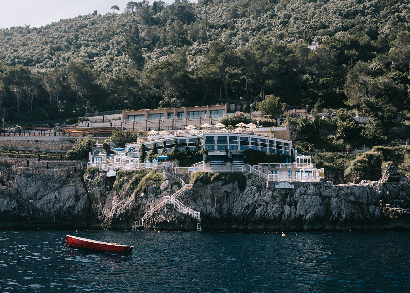 A cliffside restaurant with multiple terraces adorned with umbrellas is nestled among dense green trees. Below, the Red Boat by Francesca Owen floats on the calm waters near the rocky shore. Terraced walkways and stairs connect different levels of the venue, creating a scene worthy of fine art prints available for worldwide shipping.