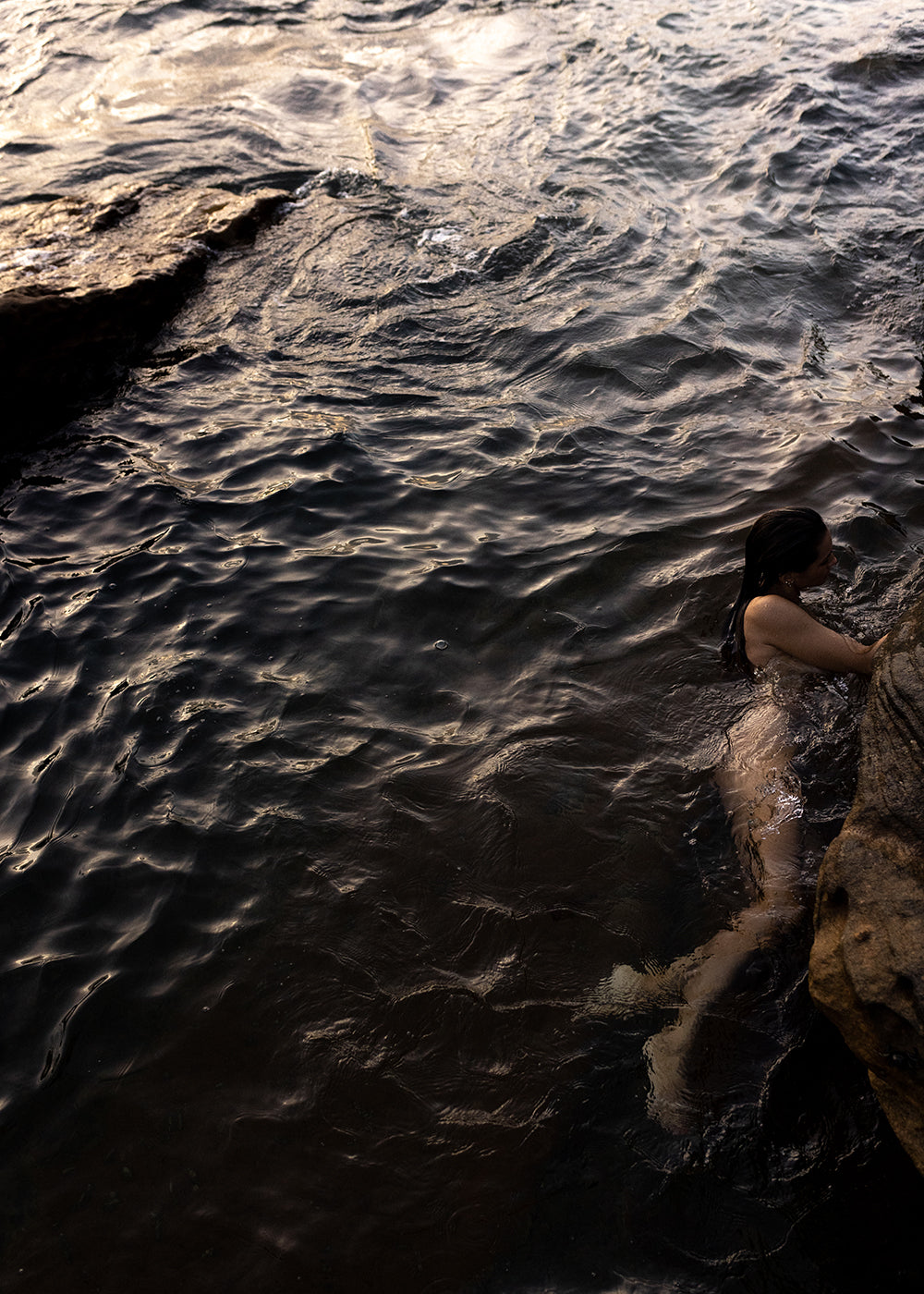 A person with long dark hair swims towards a rocky shoreline in rippling, dark water. The light reflects off the water's surface, creating a play of shadows and highlights, reminiscent of *Sunrise Swim #5* by Francesca Owen, crafted with fine art paper and archival pigments. The person's hand reaches out to grab the rock.