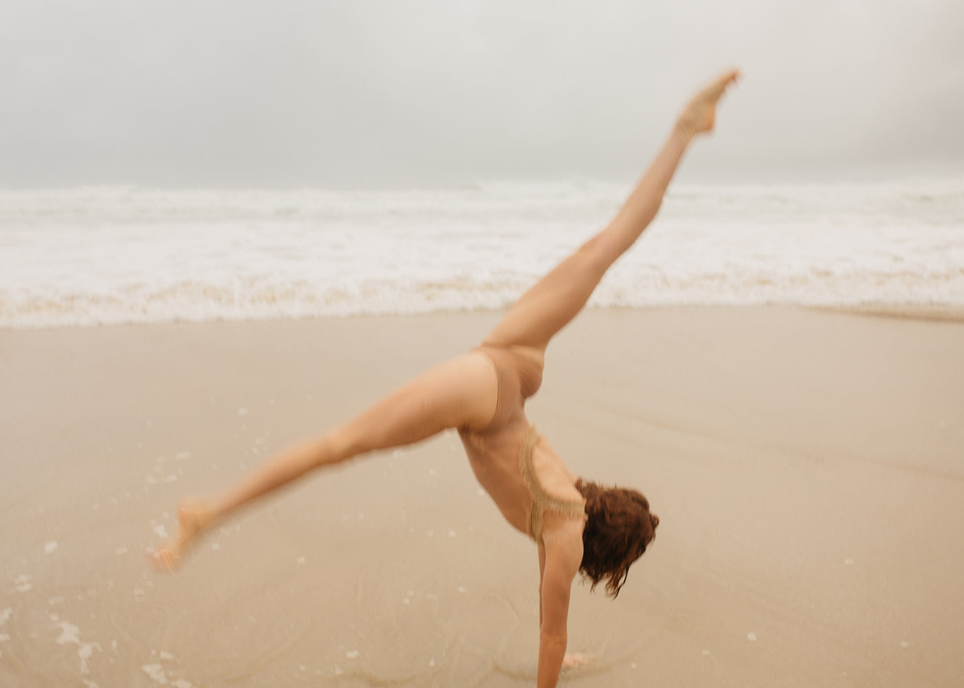 A person in a swimsuit performs a cartwheel on a sandy beach with the ocean in the background. The sky is overcast, creating a serene and peaceful atmosphere, evocative of Francesca Owen's Red Head Series #4, meticulously captured on fine art paper with archival pigments.